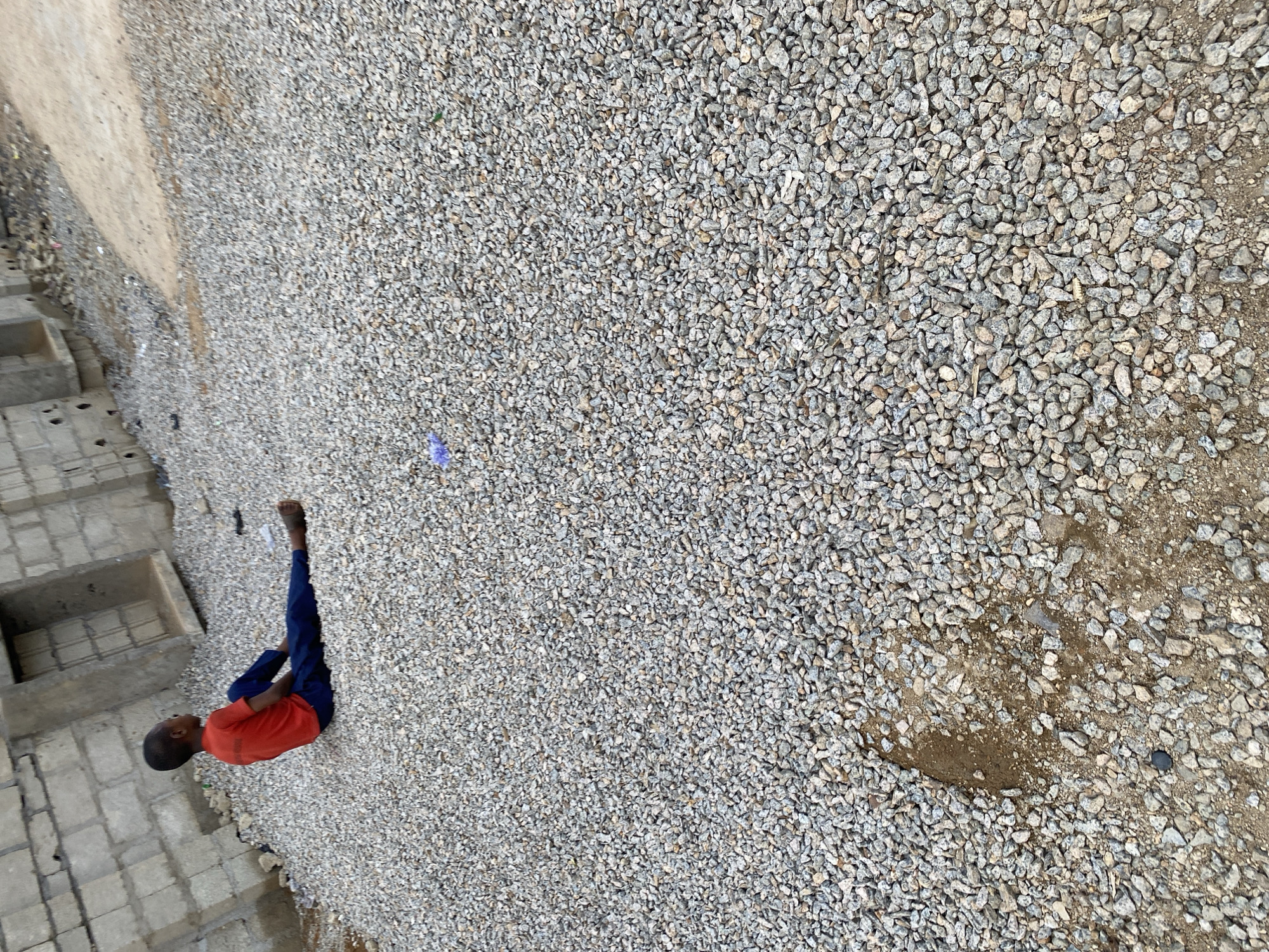In Jos, Nigeria, a young boy in a red shirt and blue trousers is seated on a surface covered in gravel. He is positioned on the right side of the frame, facing left. The background reveals a partially constructed building with grey cinder blocks. The ground consists of a mix of gravel and dirt.