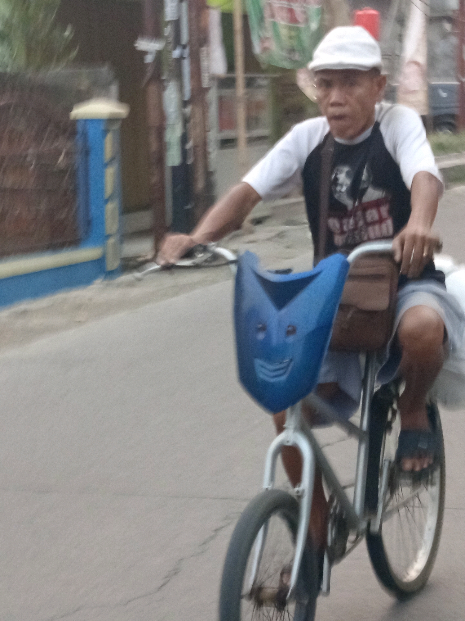 An adult male, appearing to be of Indonesian descent, rides a bicycle on a paved road in Karawang, Indonesia. He is wearing a white cap, a white t-shirt with black sleeves, and a dark t-shirt underneath with a graphic design. He is also wearing grey shorts and dark sandals. The bicycle has a blue, mask-like fairing attached to the handlebars, which features a smiley face design. A brown bag is attached to the handlebars behind the fairing. The man's hands are on the handlebars, and his feet are on the pedals. The background shows a blurry residential area with a fence and some vegetation. The lighting suggests it is daytime, possibly late afternoon.