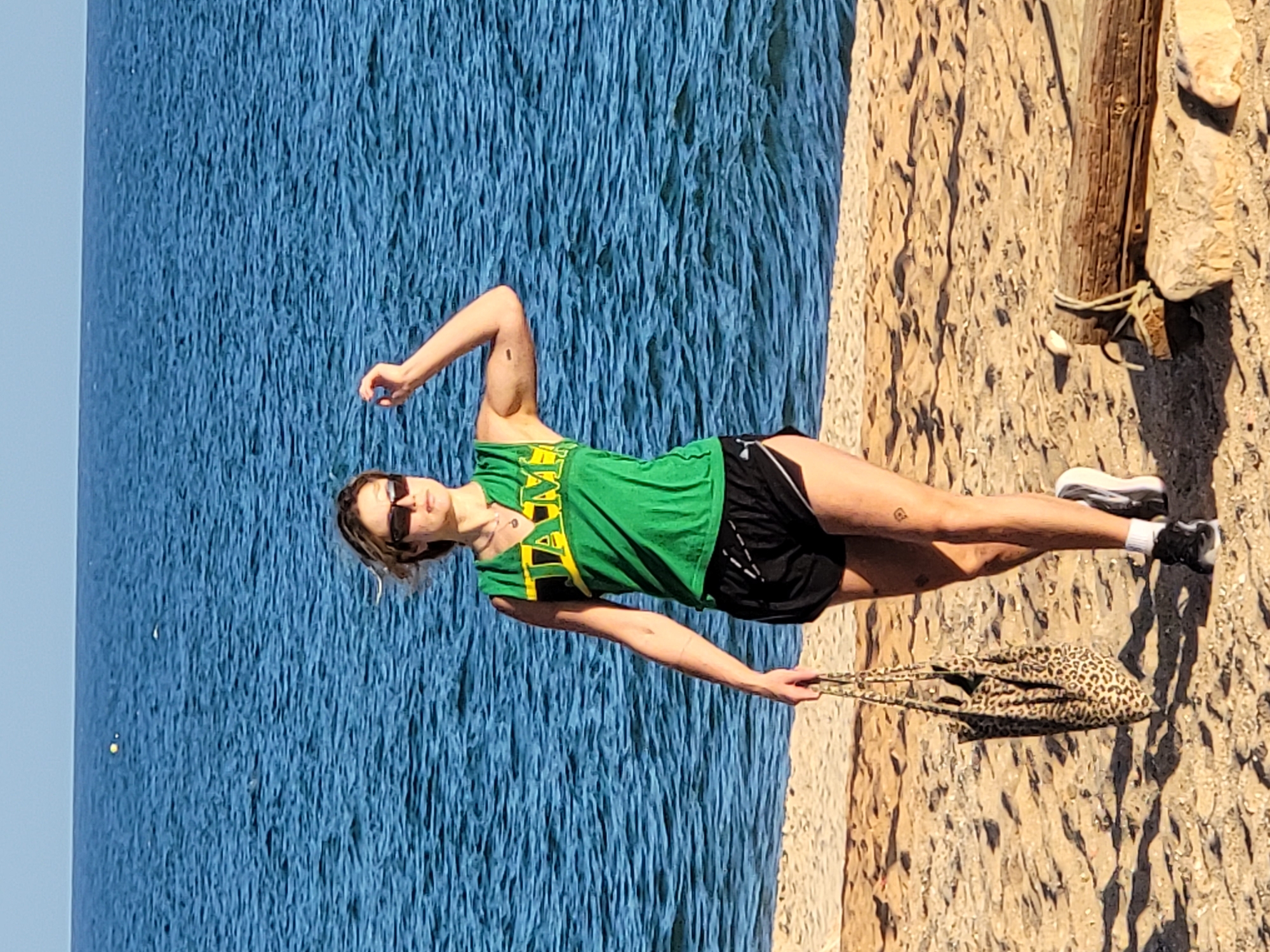 A woman walks along a sandy beach next to the ocean. She is wearing a green tank top with yellow trim and black athletic shorts. She has on sunglasses and is carrying a leopard-print tote bag. Her right arm is raised, and her left hand holds the bag. The ocean is blue and wavy. The sky is clear and blue. A piece of driftwood rests on the sand in the foreground. The location is Quarteira, Portugal.
