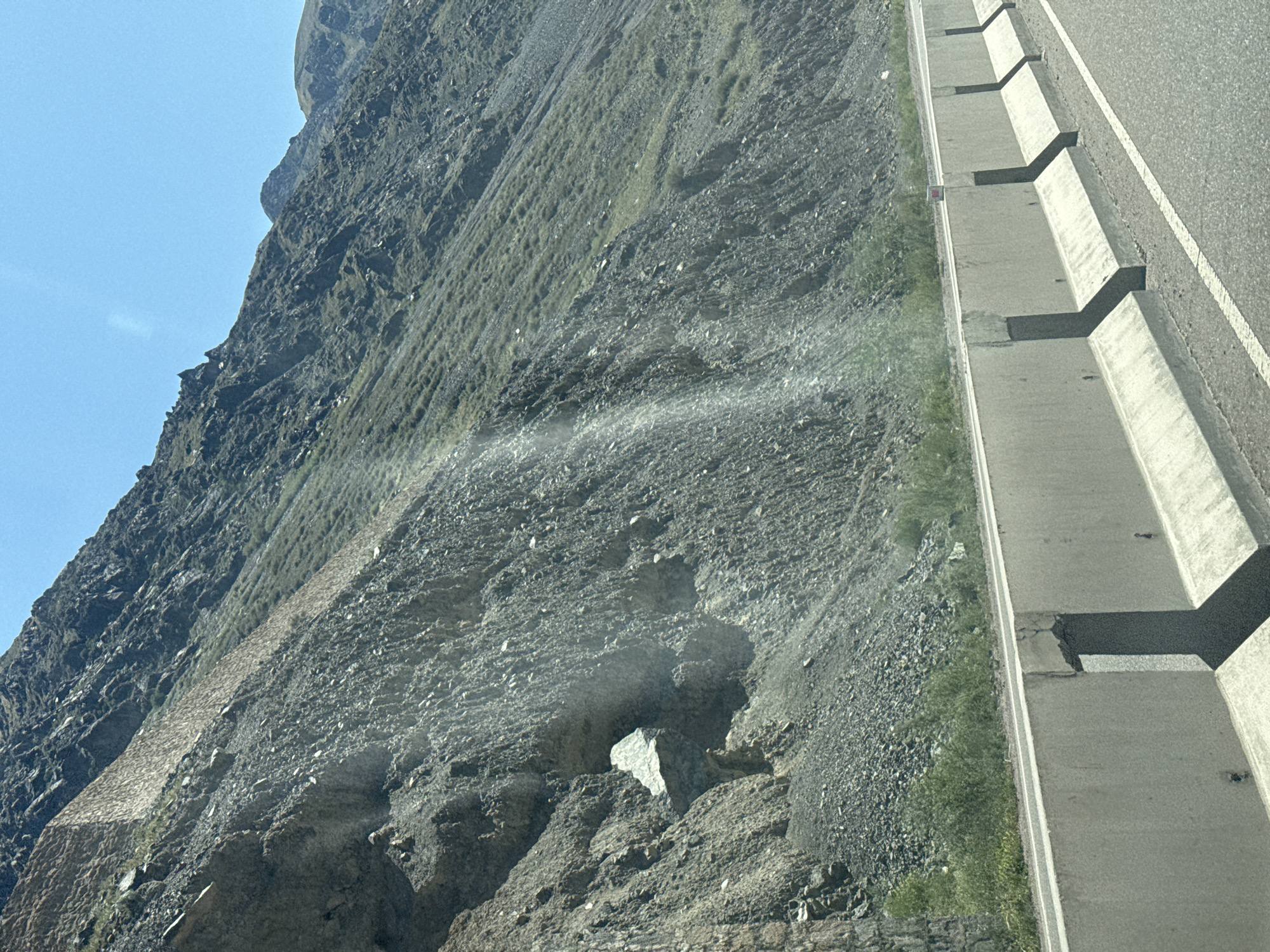The image shows a barren, rocky mountainside adjacent to a paved road with a concrete barrier. The mountainside is covered in loose scree and sparse vegetation. There is a visible rock formation with an opening on the mountainside, and a bright beam of sunlight illuminates a section of the slope. The sky is clear and blue. The road appears to be an elevated highway or a major road. The overall impression is one of a rugged, remote landscape. The scene is devoid of any human presence. The exact location within Kyrgyzstan is not specified.