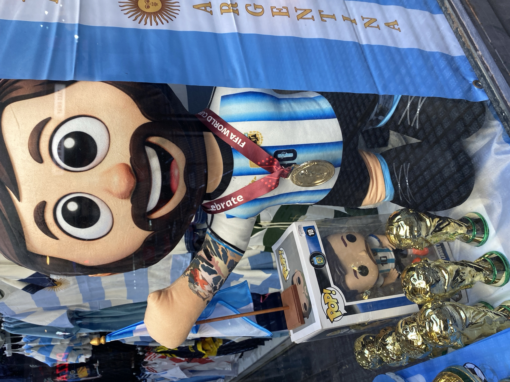 The image depicts a shop window display in Buenos Aires, Argentina, featuring Argentina national football team memorabilia. The display includes an Argentina flag, a large plush doll resembling Lionel Messi wearing a blue and white jersey with a FIFA World Cup lanyard, a Lionel Messi Funko Pop figure, and miniature gold-colored replica World Cup trophies.