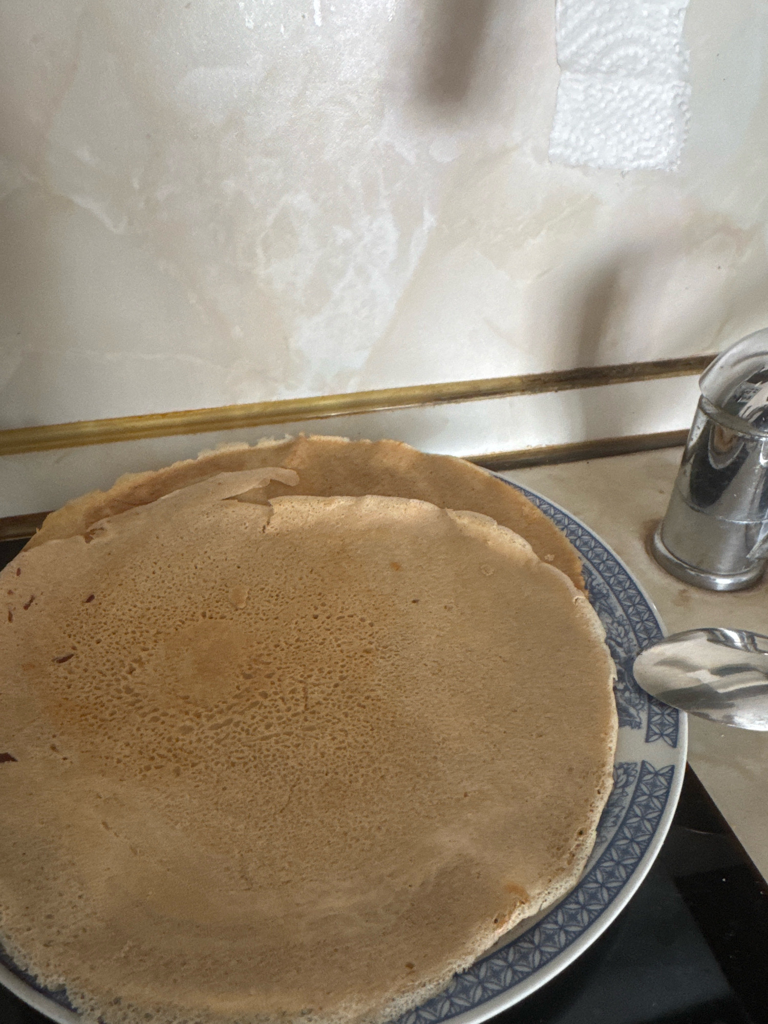 A close-up, slightly angled shot captures a plate of crepes on a countertop. The crepes are a light golden brown with a slightly textured surface, and they are stacked on top of each other. The plate has a blue and white decorative rim. To the right of the plate, a chrome salt shaker is visible, and a portion of a spoon rests on the rim of the plate. The background consists of a tiled wall with a paper towel fragment attached. The scene appears to be set in a kitchen environment in Sofia, Bulgaria.