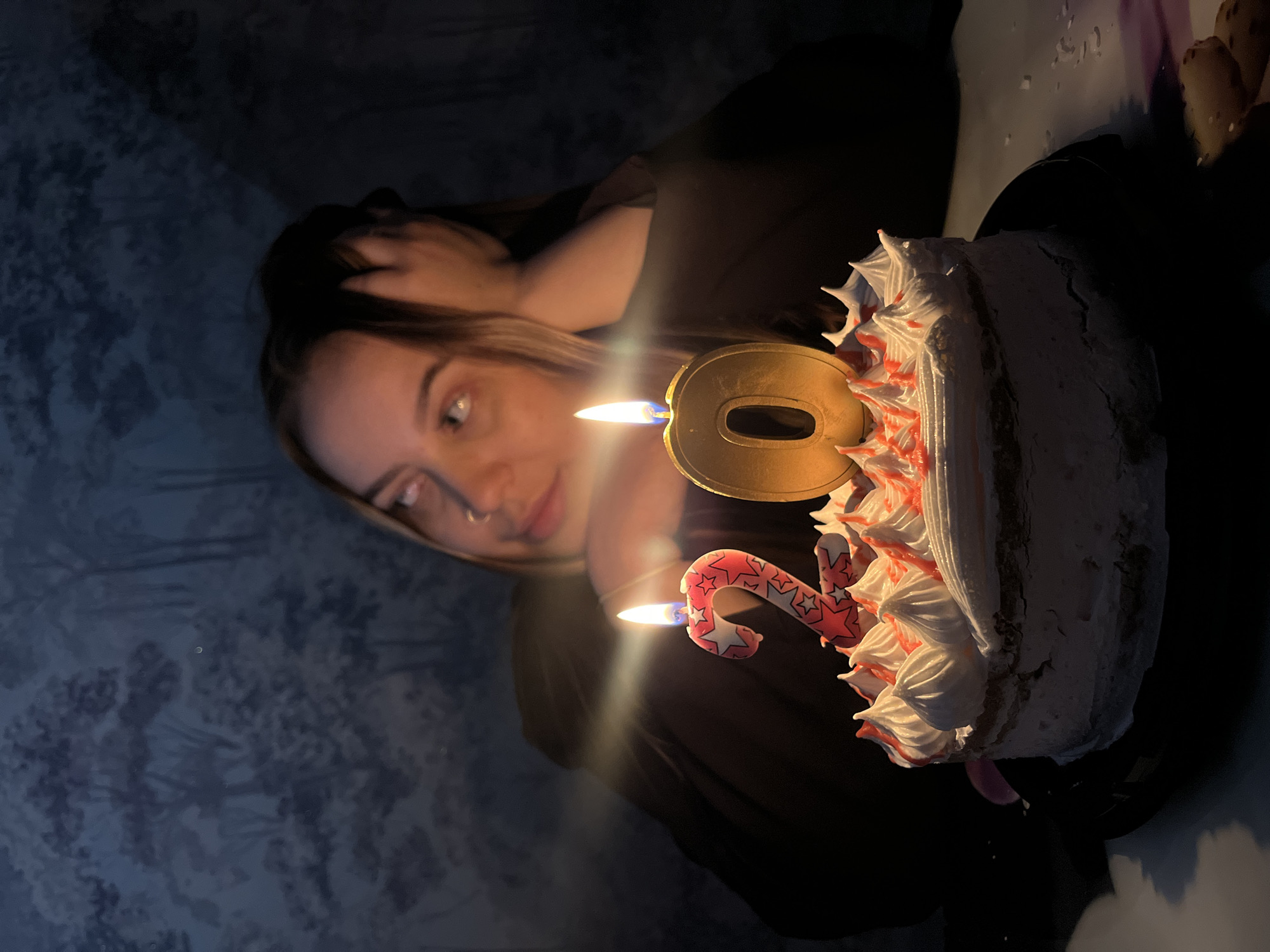 A young woman, likely in her late teens or early twenties, is captured in a dimly lit indoor setting. She is leaning on her hand, gazing upwards with a thoughtful expression, while resting her head on her arm. In front of her, a frosted cake with white icing and red accents is adorned with two lit birthday candles. The candles are shaped as the numbers '2' and '0', indicating a twentieth birthday. The background features wallpaper with a dark, arboreal pattern. The scene suggests a personal celebration, possibly in a residential environment in Novopokrovskaya, Russia, given the provided context.