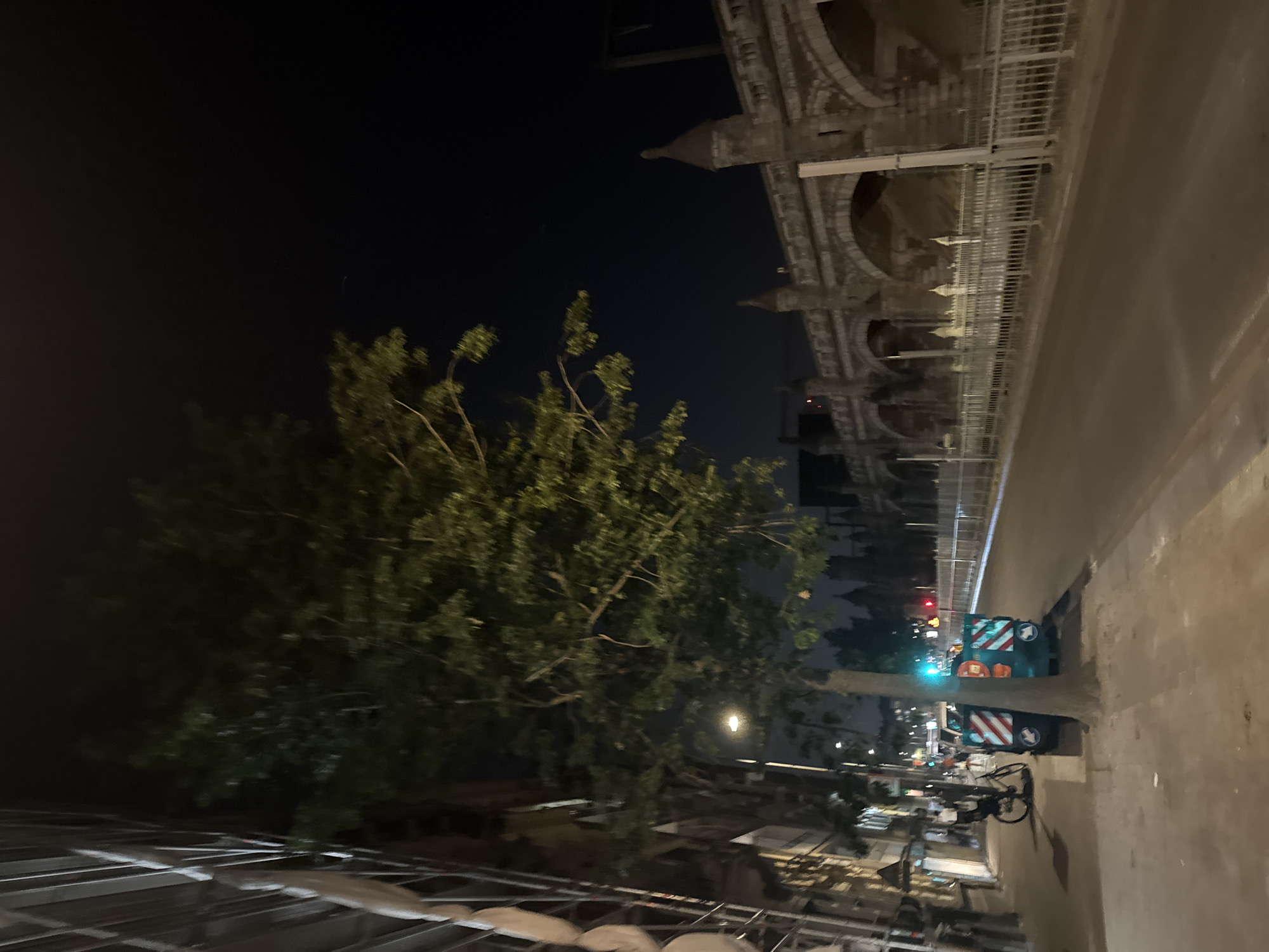 The image depicts a nighttime scene on a city street. On the left side of the image, a building is under construction, with scaffolding visible. A tree with green leaves stands in the foreground, partially obscuring a street lamp. Further down the sidewalk, there is a bicycle parked next to a green and white striped trash bin. Across the street, a long, arched stone bridge dominates the background, with a metal fence in front of it. The sky is dark, indicating it is nighttime. The street appears to be empty of moving vehicles, with only a few distant lights visible.