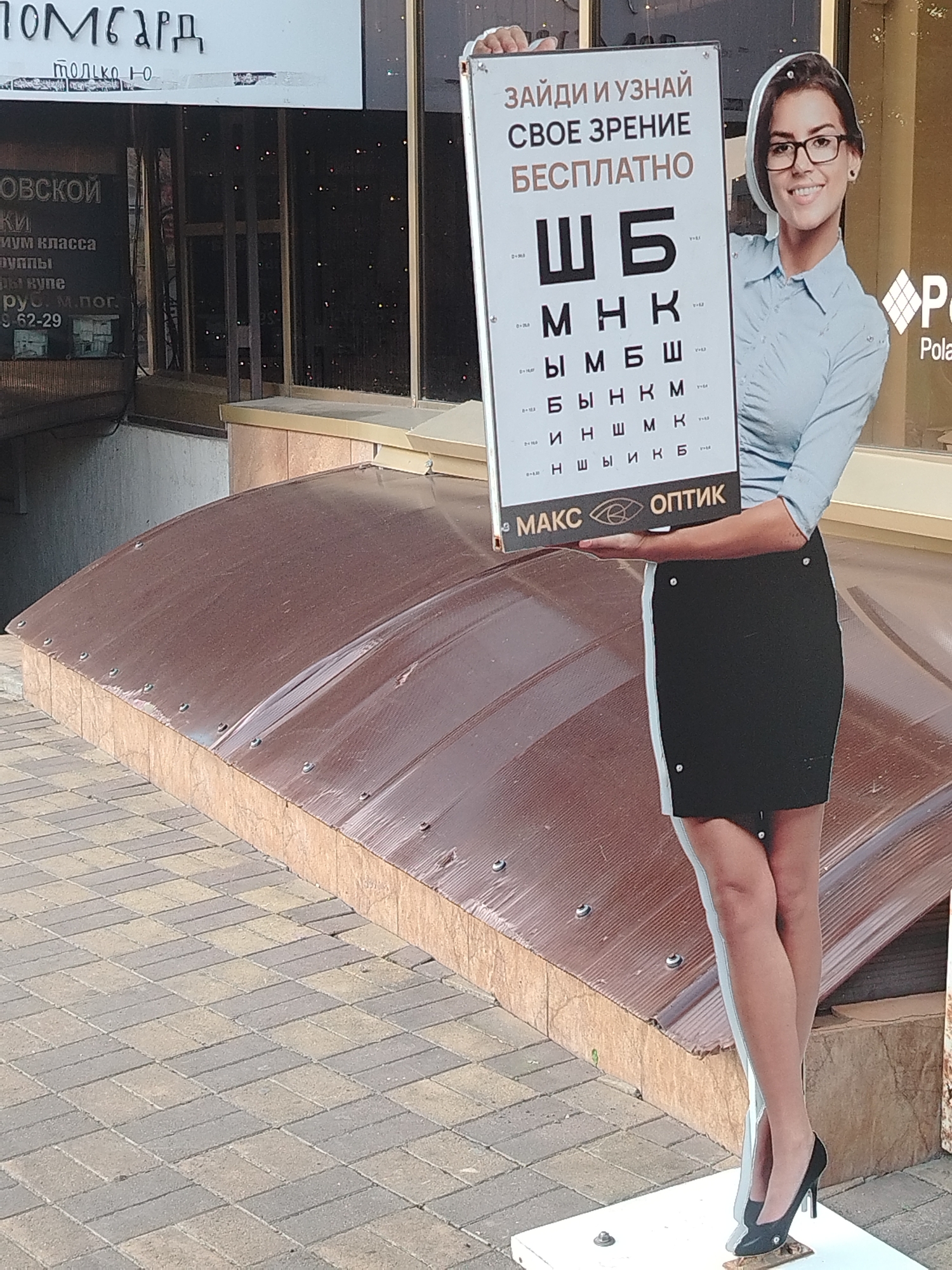 The image shows a cutout of a woman holding an eye chart. The woman is wearing glasses and a light blue collared shirt with a black skirt. The eye chart has Russian text and letters on it, indicating it's for vision testing. The background appears to be a storefront with signage, and part of a brown awning is visible in the foreground. The ground is paved with light and dark gray interlocking bricks. The signage in the background appears to be in Russian, with "ПОМБАРД" and "ТОЛЬКО Ю" visible at the top. Below that, "ОВСКОЙ", "ки", "им класса", "уппы", "ы купе", "рус. М.Пог.", and "9 62-29" are visible. Another sign has the words "ЗАЙДИ И УЗНАЙ", "СВОЕ ЗРЕНИЕ", "БЕСПЛАТНО", followed by letters and the text "МАКС ОПТИК". Based on the text and context, this appears to be an advertisement for an optician's shop, likely located in Russia, possibly Krasnodar given previous associations with similar imagery.