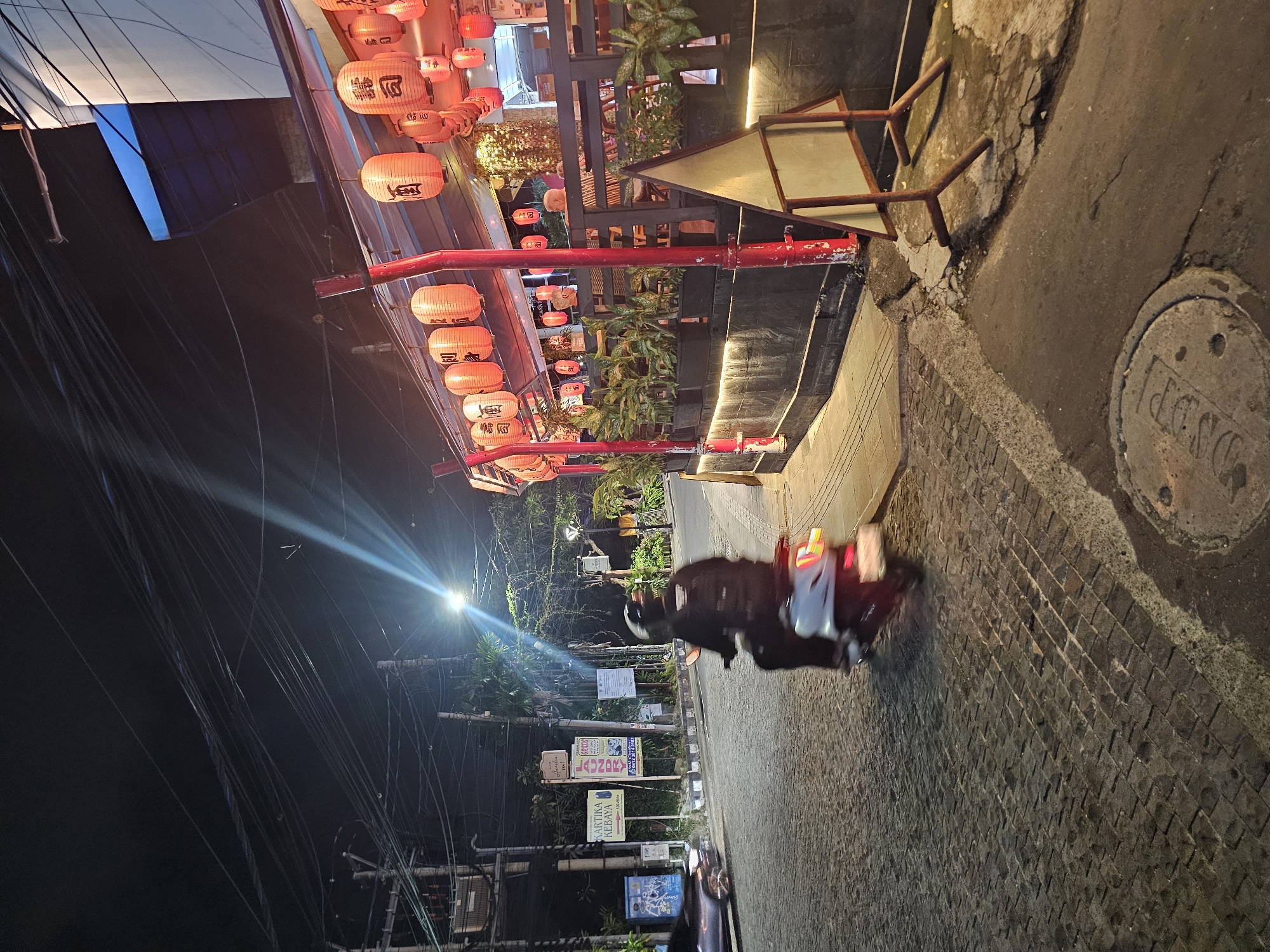At night, a person rides a scooter on a narrow brick road in Denpasar, Indonesia. Overhead, rows of illuminated red lanterns decorate an establishment with dark wooden architectural elements and potted greenery. A street lamp illuminates the scene. Signs advertising various businesses line the opposite side of the street from the lanterns. Overhead power lines run along the left side of the frame.