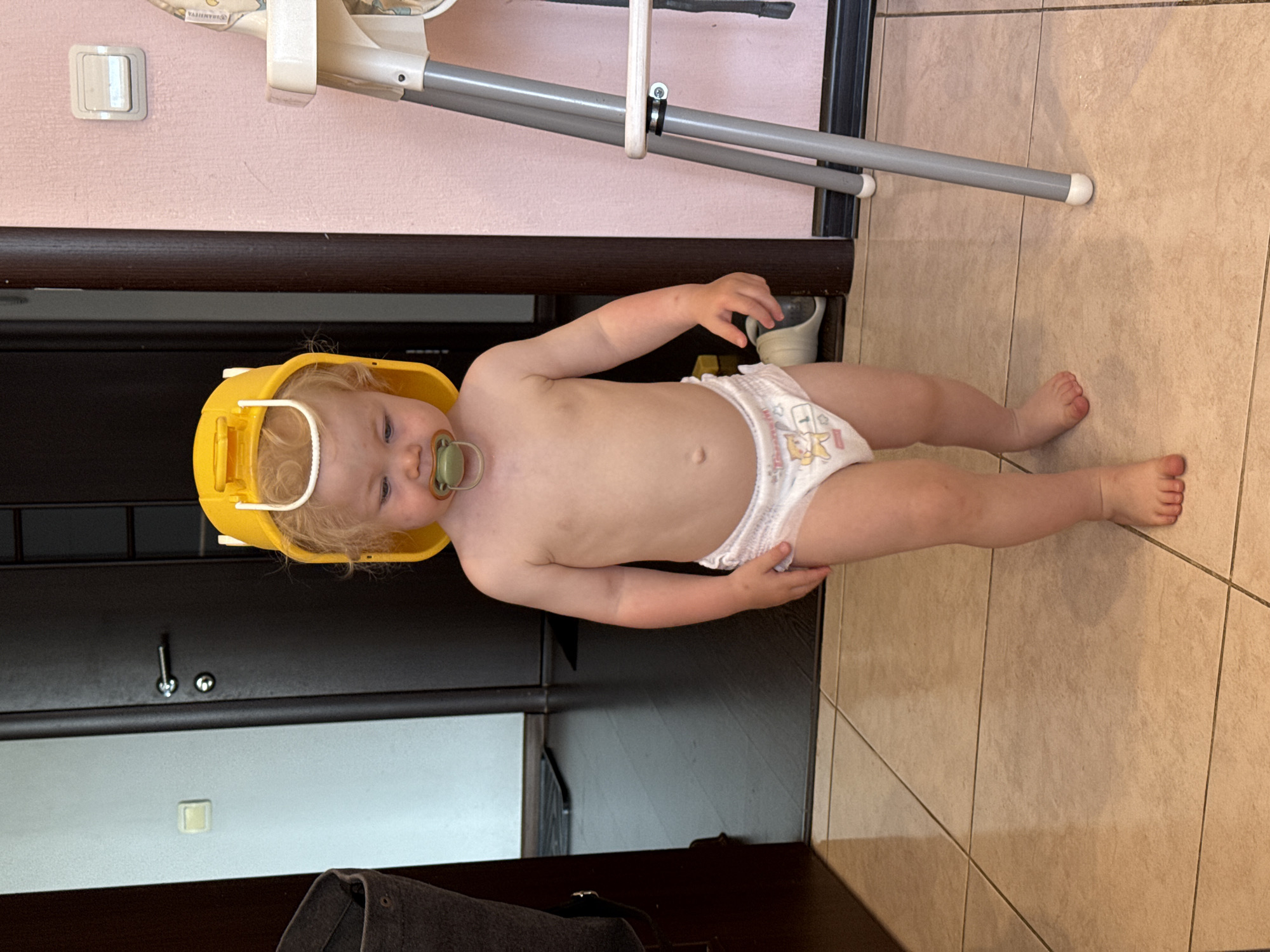 A toddler wearing a yellow plastic bucket as a hat and a diaper stands on a tiled floor indoors. The child has a pacifier in its mouth and is looking down. To the left of the child is a dark brown doorframe and a portion of a dark closet or piece of furniture. To the right is a high chair with gray legs and a white seat, and a pink wall. The floor is tiled in a beige, square pattern. The lighting suggests it is daytime. The image does not provide any information about its specific location in the city of Vladimir, Russia.