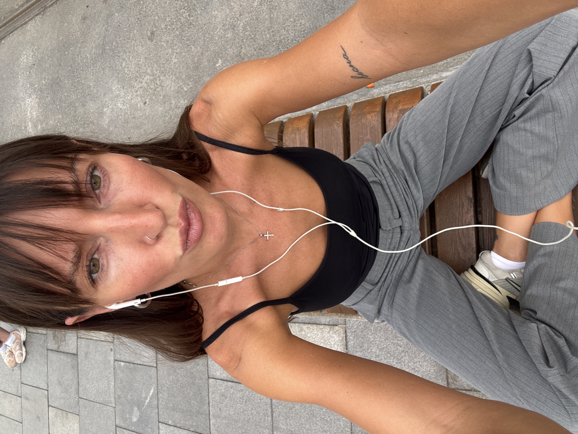 A young woman is sitting on a wooden bench, facing the camera at a close range. She has long, dark brown hair with bangs, and is wearing a black tank top, grey pinstripe pants, and white sneakers. White earbuds are in her ears, with the wire visible. She has a delicate silver necklace with a cross pendant. On her left arm, there is a tattoo. She appears relaxed, with a slight smile and her eyes are looking towards the camera. The scene is outdoors, with a paved ground and the wooden bench. The lighting suggests it is daytime. The setting appears to be a public outdoor space in Moscow, Russia.