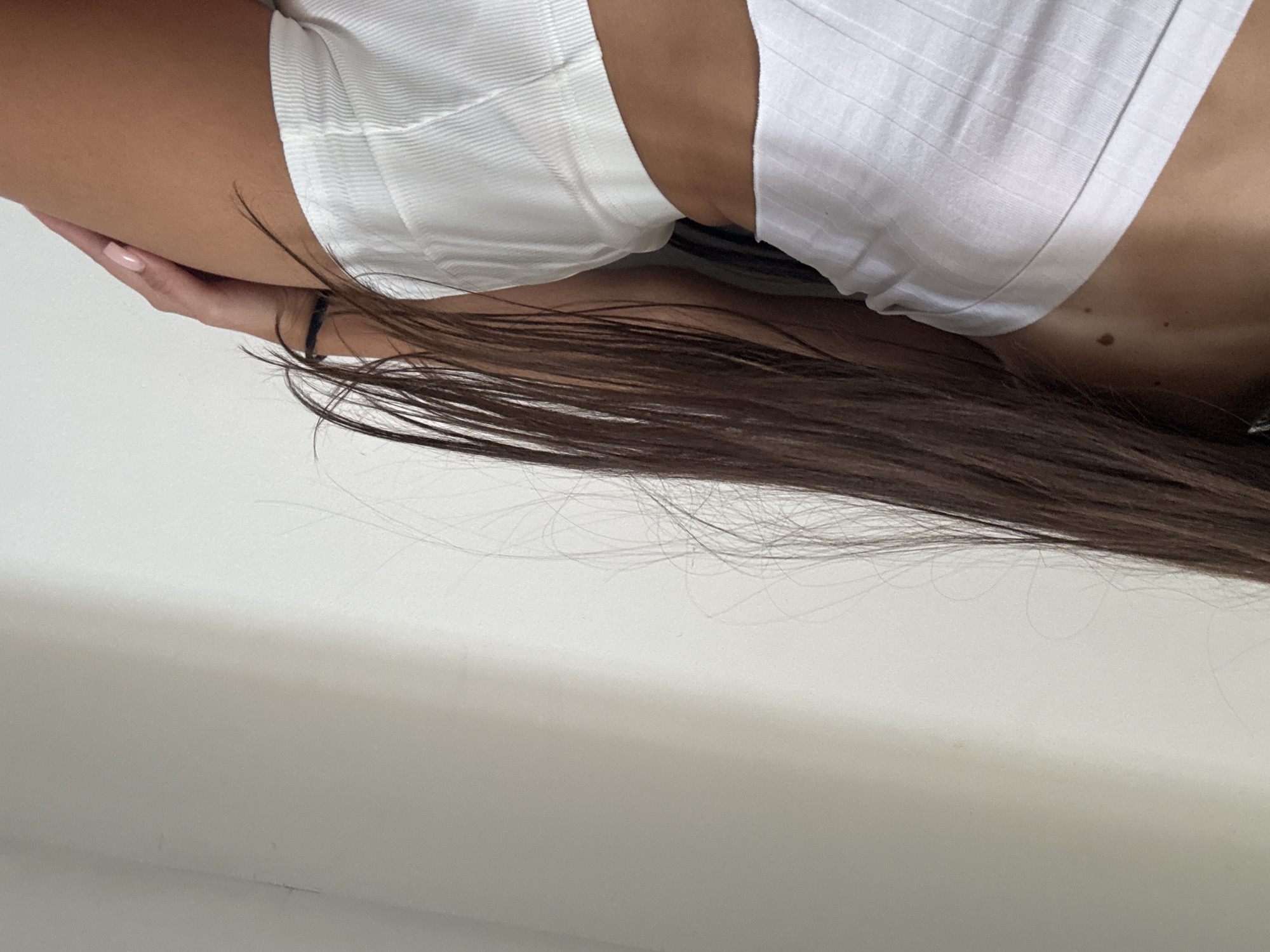 A close-up, side-view shot shows the mid-section of a person with tanned skin. They are wearing a white ribbed tube top and matching white ribbed shorts. Long, dark brown hair cascades down their back, partially obscuring their right side. Their right hand, with light pink nail polish, rests on their thigh. The background is a plain, off-white wall, which appears to be indoors. There is no discernible information to place this image in Chișinău, Moldova.