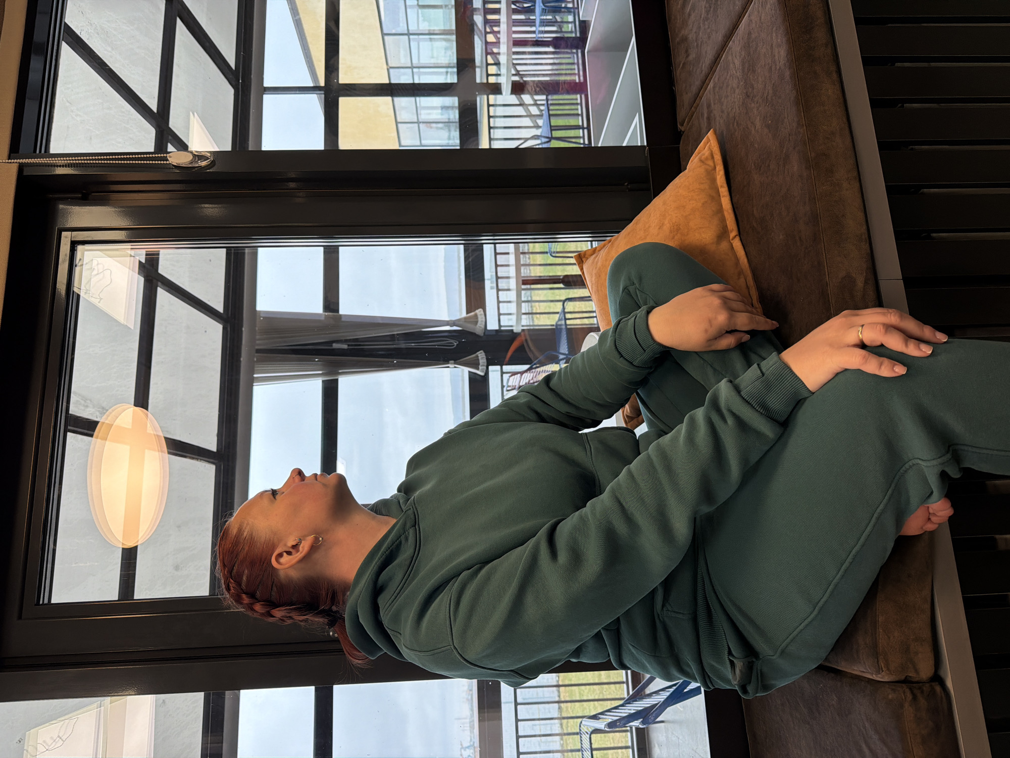 A woman with red hair braided to the side is sitting on a brown cushioned bench in what appears to be an indoor space. She is wearing a green sweatsuit and is leaning against a brown pillow. Behind her is a large window with black frames, reflecting the exterior view including buildings, possibly of a modern architectural style, and a glimpse of what appears to be a landscape. A hanging oval light fixture is also visible near the window. Based on the prompt, the location is Chaikovsky, Russia. The woman's hands are resting on her knee.