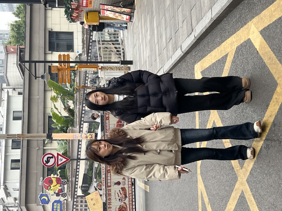 This image depicts an urban street scene in Liangjiang, China, under an overcast sky. Two East Asian females are walking side-by-side, moving from the background towards the foreground.
The female on the left has long, dark brown hair and is wearing a light beige or tan puffer jacket with a brown fur-trimmed hood, dark blue flared jeans, and light-colored sneakers. She holds a white smartphone in her left hand, and her right arm is linked with the left arm of the adjacent female. Her facial expression is neutral.
The female on the right has long, straight black hair and is wearing a black puffer jacket, dark-colored flared pants, and light brown or tan platform shoes. A dark shoulder bag is visible across her body under her jacket. Her facial expression is neutral.
In the immediate foreground, a yellow cross-hatch pattern is painted on the dark asphalt road. To the right of the individuals, a gray paved sidewalk is present, leading towards the background.
The background features multiple urban elements. On the left, a tall, narrow pole stands near a large mural or billboard designed with a film strip motif. The mural includes a yellow cartoon character on a scooter, a blue crescent moon, a red circular 'yield' sign with the Chinese character "让" (ràng), and various other images and Chinese text, some appearing to be menu items. Behind this, multi-story buildings with windows are visible.
On the right side of the background, another pole supports a street lamp and a yellow directional signpost with multiple arrows and Chinese text. A white metal railing is situated along the edge of the sidewalk, and a bright yellow rectangular box, possibly a mailbox or utility box, is mounted on the sidewalk further back. Green foliage, resembling banana leaves, protrudes above the railing between the buildings. Additional indistinct figures are visible further down the street in the background.