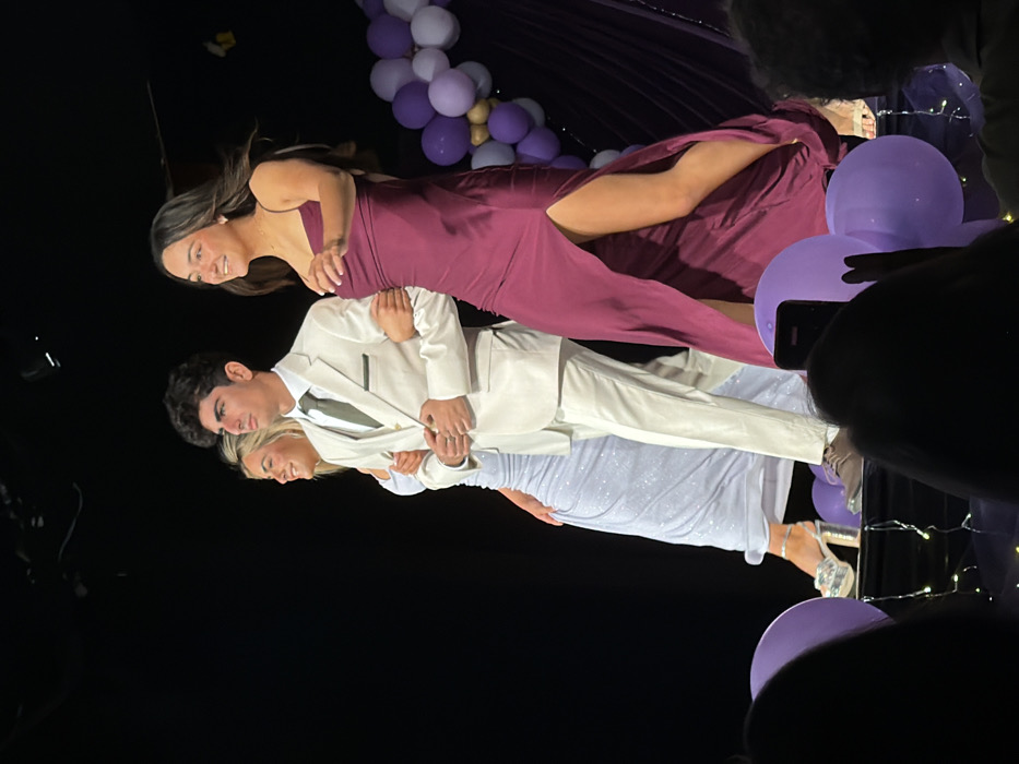 The image captures three young adults walking left to right across a spotlighted stage indoors, located within Palos Verdes Estates, United States. A male with dark hair occupies the central position, dressed in a cream-colored suit, a light vest, and a dark tie. He is linked arm-in-arm with two females. To his left, a female with blonde hair wears a sparkling light blue, floor-length gown and silver platform heels; her left arm is linked with the male's left arm, and she is smiling. To his right, a female with dark hair wears a dark magenta, floor-length gown featuring a high left-side slit; her right arm is linked with the male's right arm, and she is also smiling. The background is primarily dark, with an arrangement of purple and gold balloons visible on the right side of the stage. The blurry foreground shows the heads of audience members, one of whom is holding a mobile phone, along with two large purple balloons and string lights on the floor in front of the stage.
