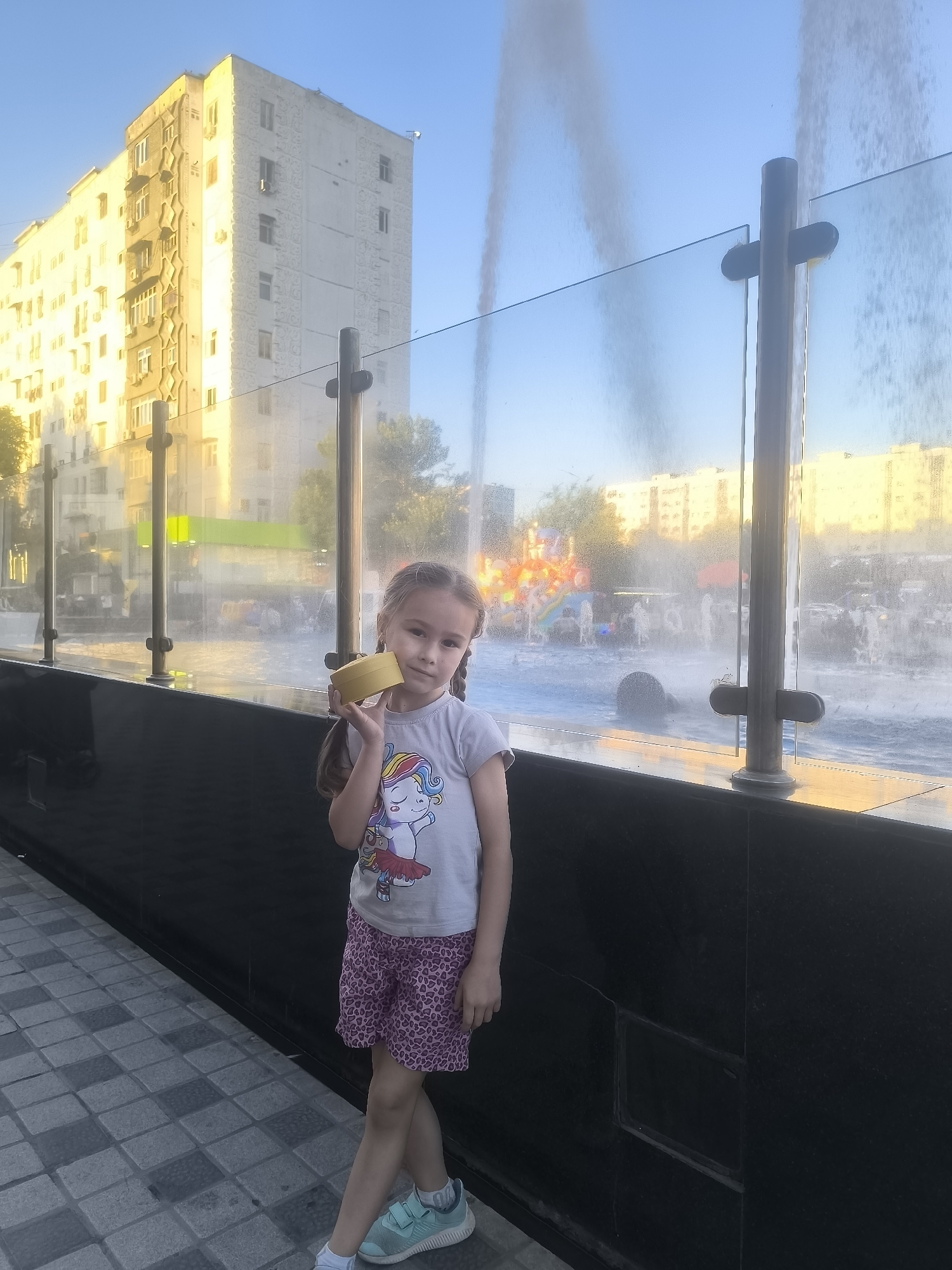 A young girl stands in front of a fountain in Tashkent, Uzbekistan. She wears a gray shirt with a unicorn design, patterned pink shorts, and blue athletic shoes. She holds a cylindrical yellow object near her face. The fountain is enclosed by a glass barrier with metal supports. In the background, a multi-story apartment building reflects the sunlight. The ground is paved with gray and white stones. The fountain water is actively spraying. Beyond the fountain, other buildings and greenery are visible.