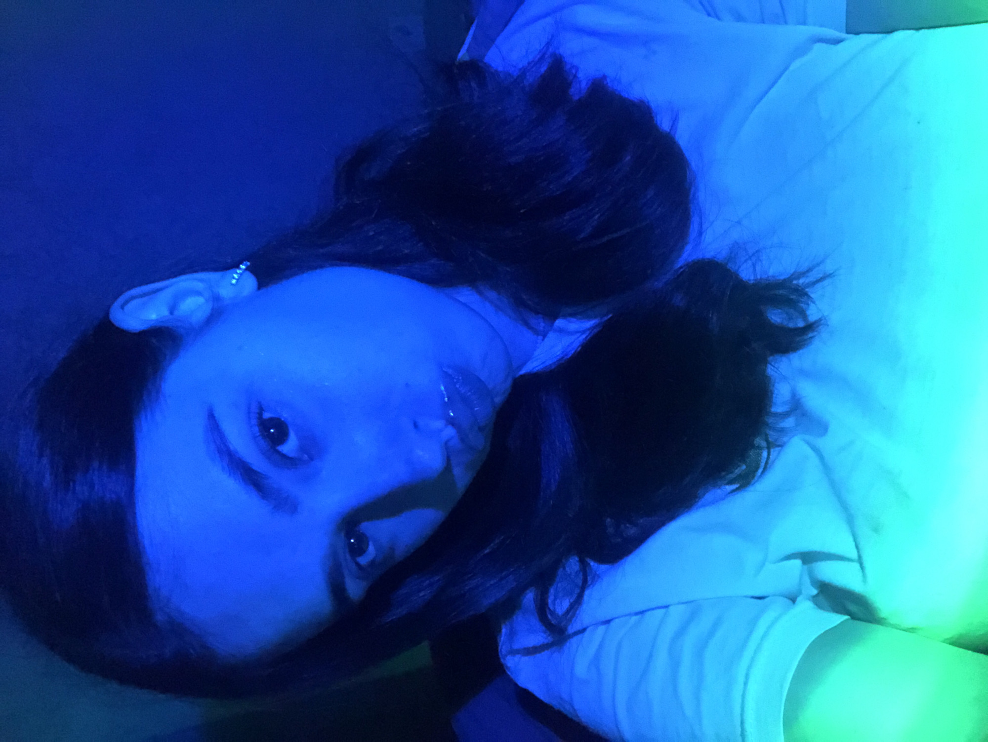 A young woman with dark hair and a white shirt poses for a selfie, illuminated by blue and green lighting. Her head is tilted to the side, and she is looking directly at the camera with her lips slightly parted. She is wearing a small hoop earring in her visible ear. The background is dark and indistinct. The image is a close-up portrait, with the focus on the woman's face and upper body. The overall impression is one of dramatic lighting and a candid moment. There is no indication of the location in Noviy, Russia.