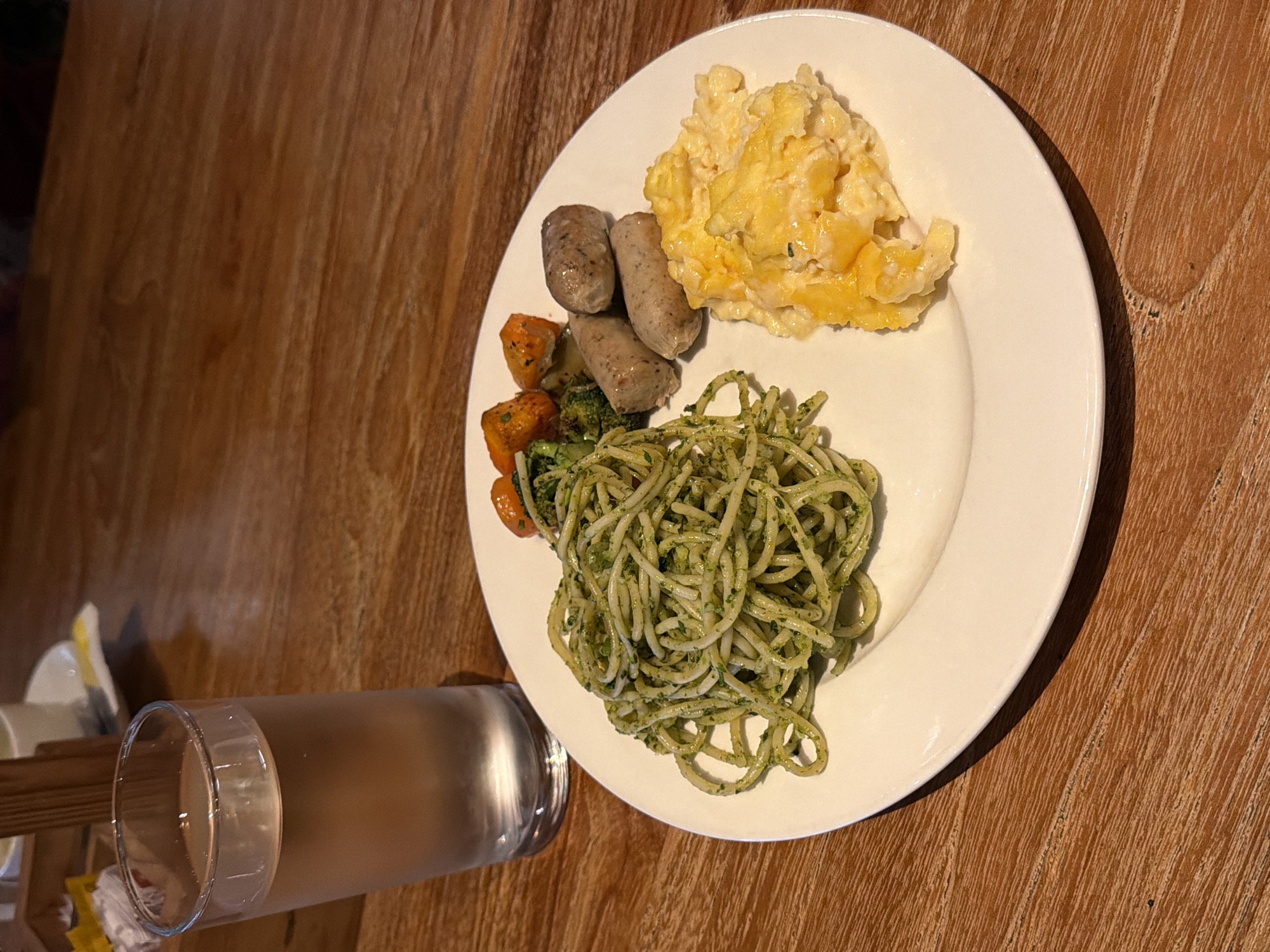 A white plate holding a breakfast meal sits on a wooden table. The plate contains a pile of green pesto spaghetti, three small sausages, a portion of scrambled eggs, and some roasted vegetables including broccoli and carrots. To the left of the plate is a tall glass of water. The background shows a blurred wooden structure and a white teacup and saucer on a doily. The scene is illuminated by natural light.