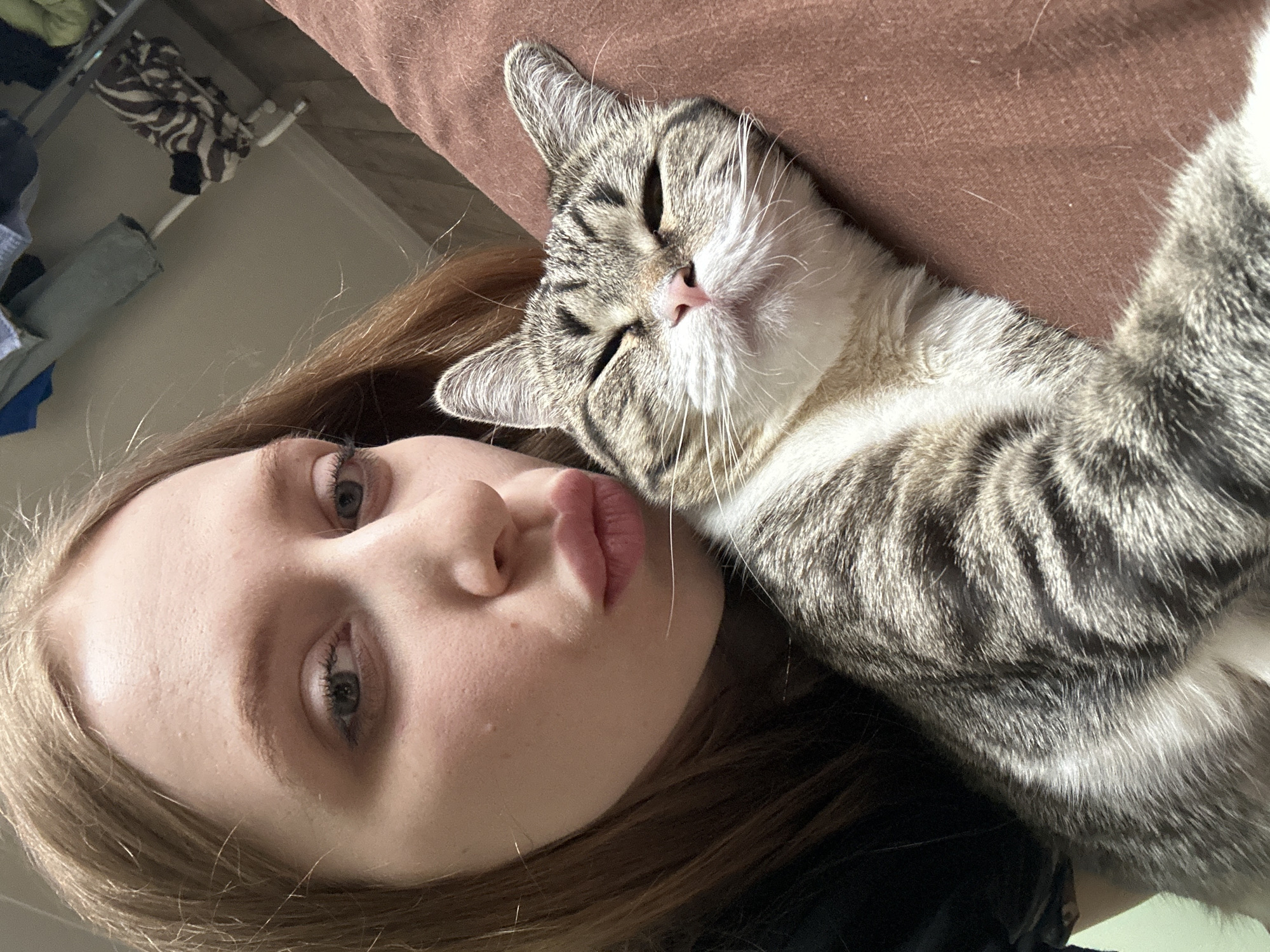 A selfie of a fair-skinned young woman with light brown hair, her face angled towards the camera, and a gray tabby cat lying on a brown surface, possibly a couch or cushion. The woman has a neutral expression, with her lips slightly pursed. The cat appears relaxed, with its eyes nearly closed. The background includes a wall and a closet with clothes hanging inside. The image's location in Chaikovsky, Russia is not discernable from the available information.