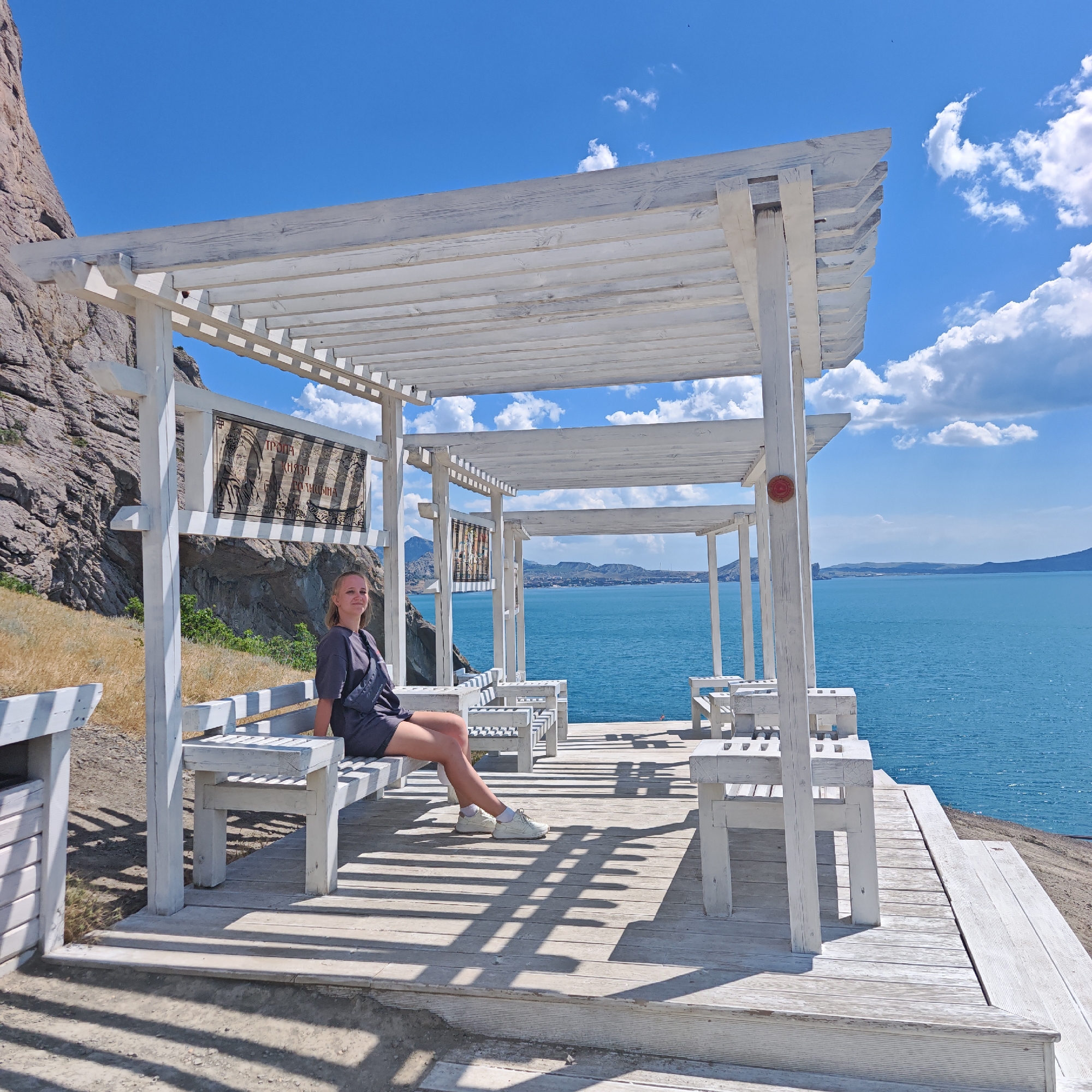 A young woman with blonde hair is sitting on a white wooden bench under a white wooden pergola. She is wearing a dark grey t-shirt and shorts, and has a grey fanny pack slung across her chest. The pergola has a slatted roof and is open on the sides, offering a view of the sea and mountains. The woman is looking towards the camera and smiling. In the background, there is a rocky mountain slope, dry grass, and the blue sea stretching to the horizon. The sky is a vibrant blue with scattered white clouds. There are several other white wooden benches and tables within the pergola area, suggesting it is a public seating area. A sign with images and text is mounted on one of the pergola's posts. The scene appears to be in a coastal, mountainous region, likely a tourist destination.