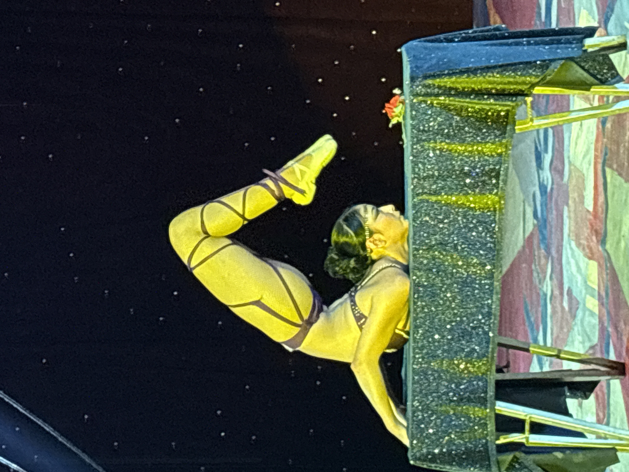 The image depicts a female contortionist performing on or near a prop resembling a building or structure. The woman is wearing a yellow and purple outfit with leg straps. Her body is arched, with her feet pointed upwards and her hands supporting her against the side of the structure. The structure appears to be decorated with glitter and gold accents. The background is dark and speckled, resembling a night sky. The location is Tejalpa, Mexico.