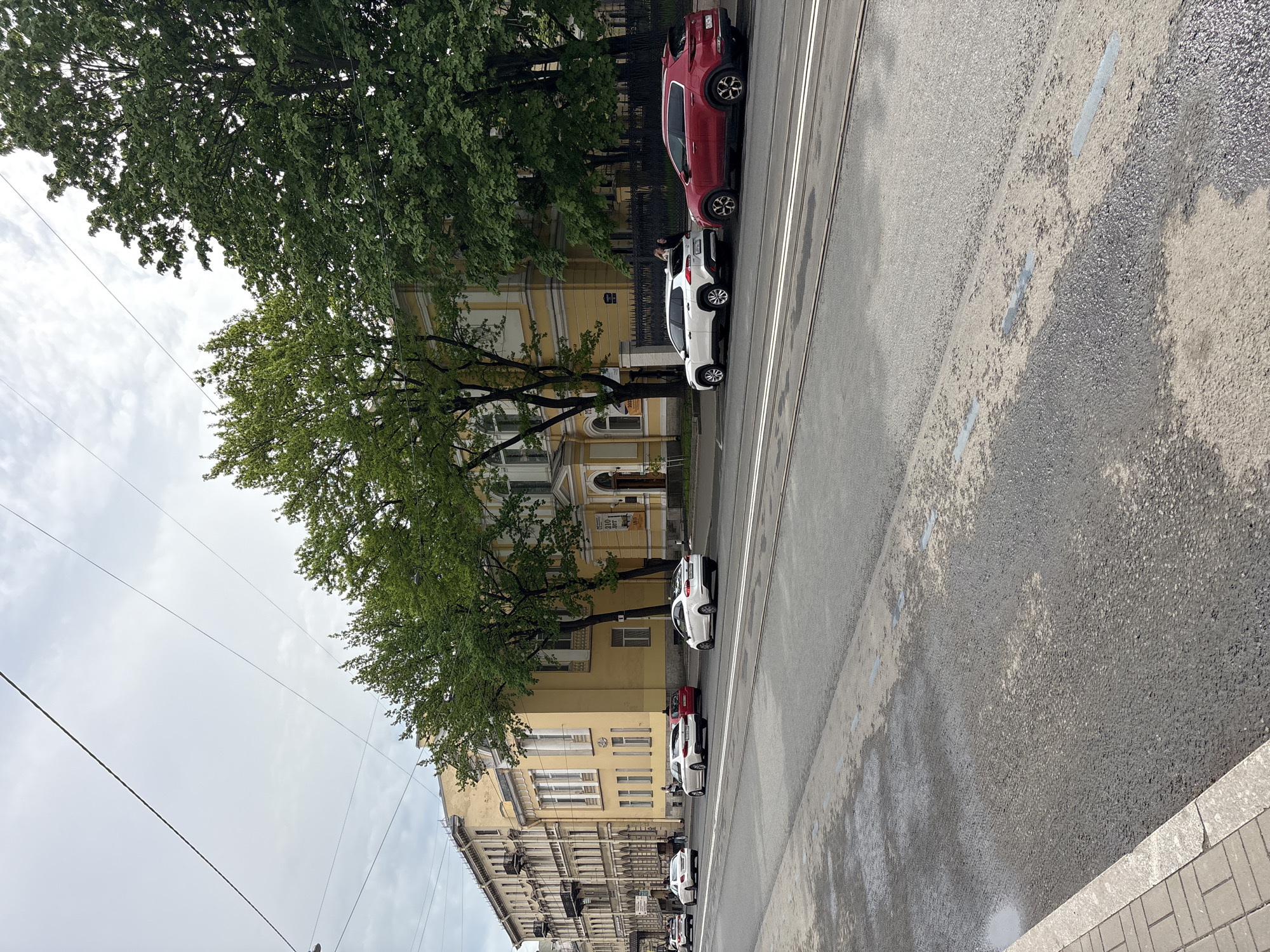 The image shows a street scene in Saint Petersburg, Russia, featuring a row of parked cars alongside a multi-story yellow building with trees lining the sidewalk. The cars, primarily white and red, are parked parallel to the street on the right side of the frame. The building is a classical style with arched windows and decorative architectural details. A cloudy sky is visible above the trees and buildings. The street appears wet, possibly from recent rain. There is evidence of tram tracks in the street.