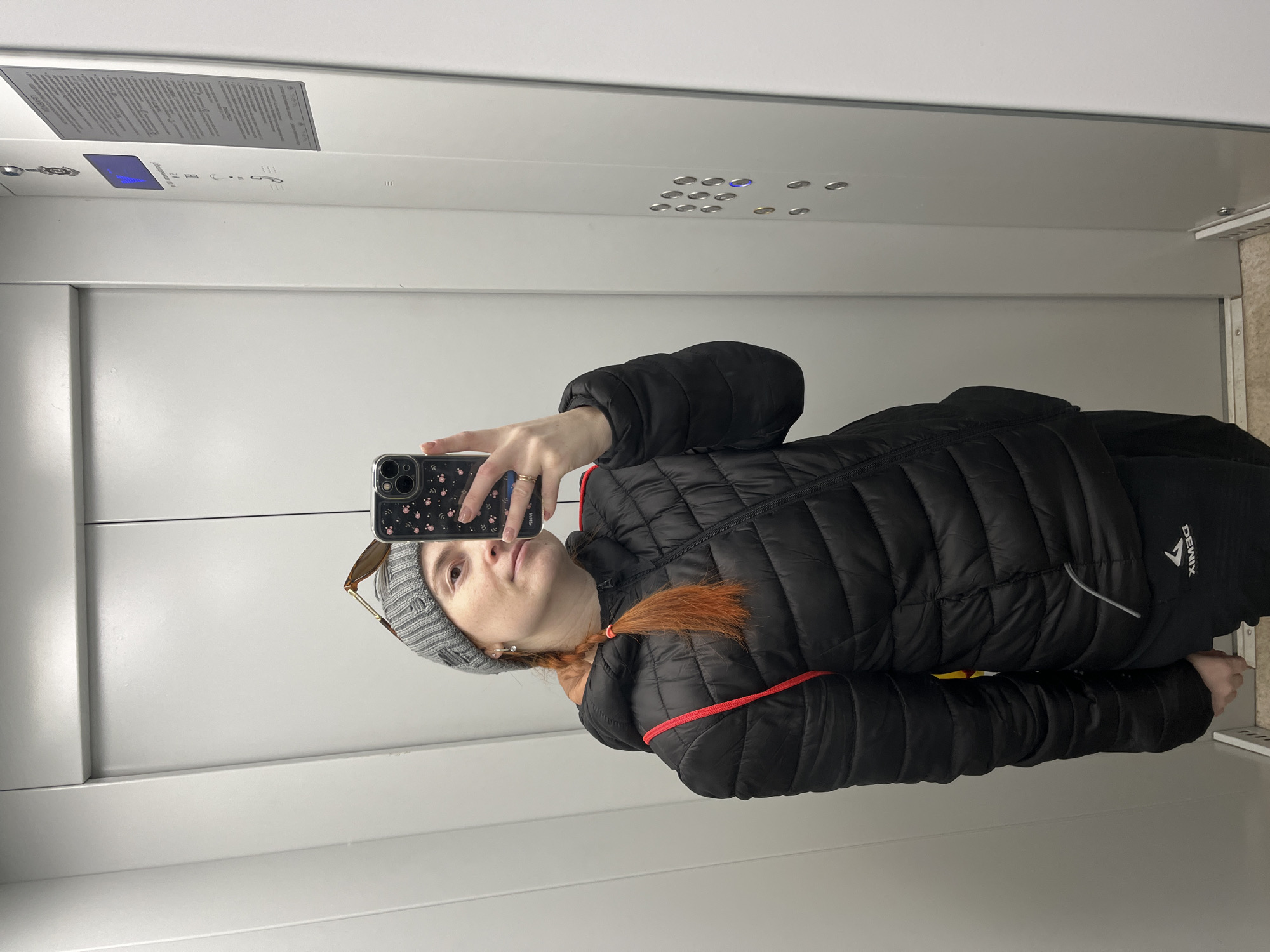 A woman stands in what appears to be an elevator, taking a selfie in the mirror. She is wearing a gray beanie, a black quilted jacket with red trim, and holding a smartphone with a floral-patterned case. The elevator has a white interior with button panels and a display screen. The location cannot be determined from the image; the city of Chaikovsky, Russia, cannot be verified.