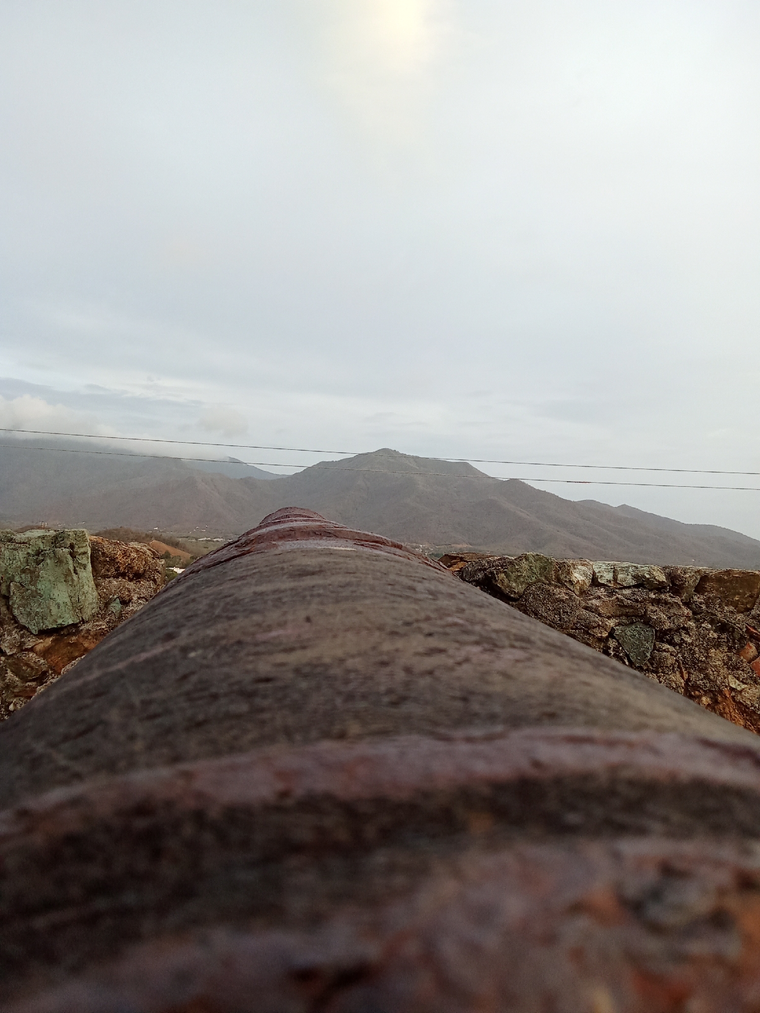 The image shows a rusted cannon barrel pointing towards a mountainous landscape under an overcast sky. The cannon is mounted on a stone structure, with visible rocks and masonry. Two power lines cut across the view of the mountains. The location is Juan Griego, Venezuela, based on the presence of a similar cannon at the Fortin de la Libertad.