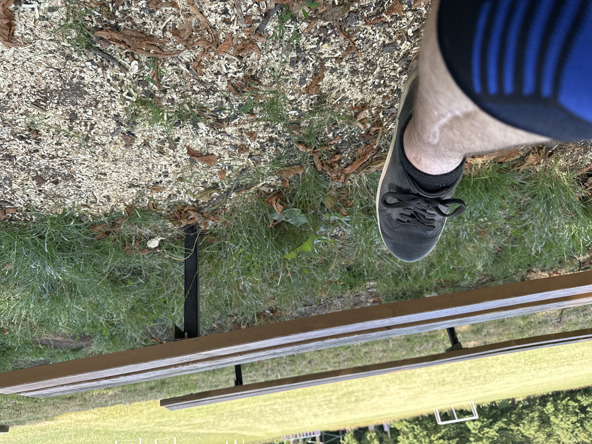 The image shows a pair of black sneakers and a person's leg wearing blue and black athletic shorts, resting on the ground. The ground is covered with wood chips, dry leaves, and some grass. In the background, two wooden benches are visible, situated in front of a grassy field. A white soccer goal is present on the field, suggesting the location is likely a sports field or park. The scene appears to be outdoors, in a setting that could be a local park or sports facility within the city of Makiv, Ukraine.