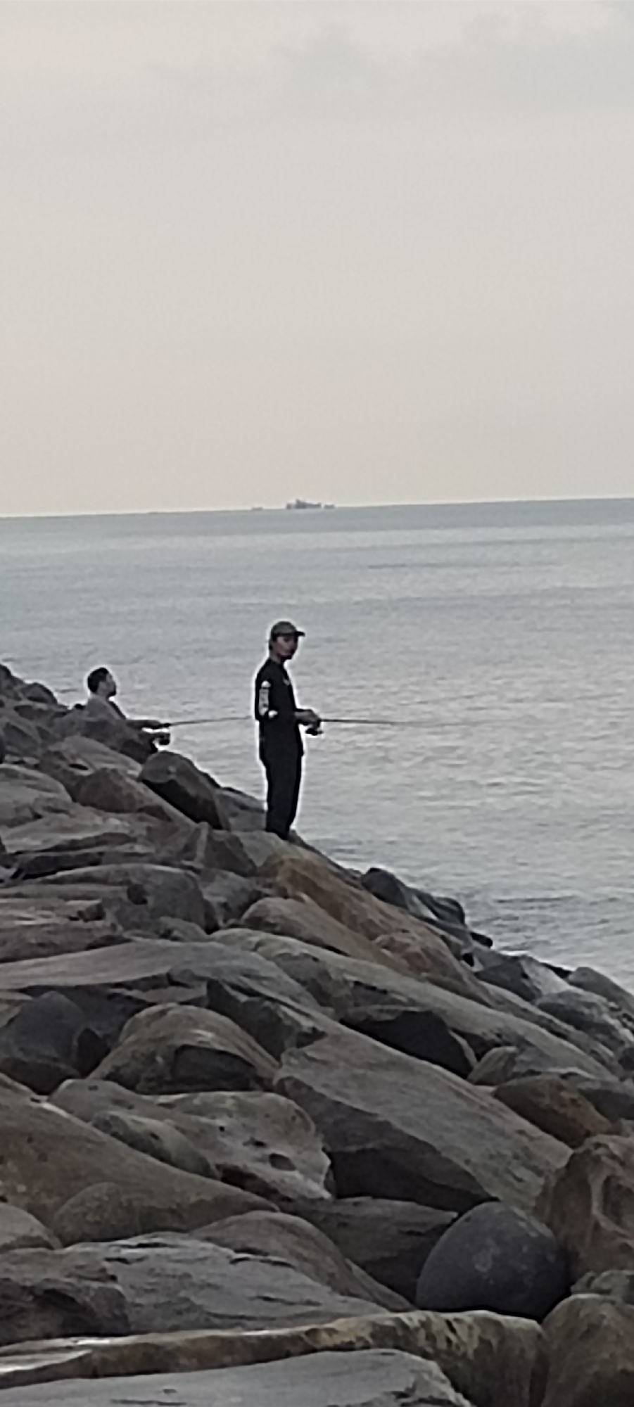 The image shows two individuals fishing from a rocky coastline. The person in the foreground is wearing a dark shirt and a baseball cap, holding a fishing rod. Behind them, another person is seated on the rocks, also engaged in fishing. The sea stretches out to the horizon, where a distant ship is visible. The sky is overcast, suggesting a cloudy or hazy day. The rocky terrain indicates a natural or man-made breakwater, typical of coastal areas. This scene is consistent with the coastline near the Port of Krueng Geukueh in Lhokseumawe, Indonesia, known for its industrial port facilities and fishing activities.