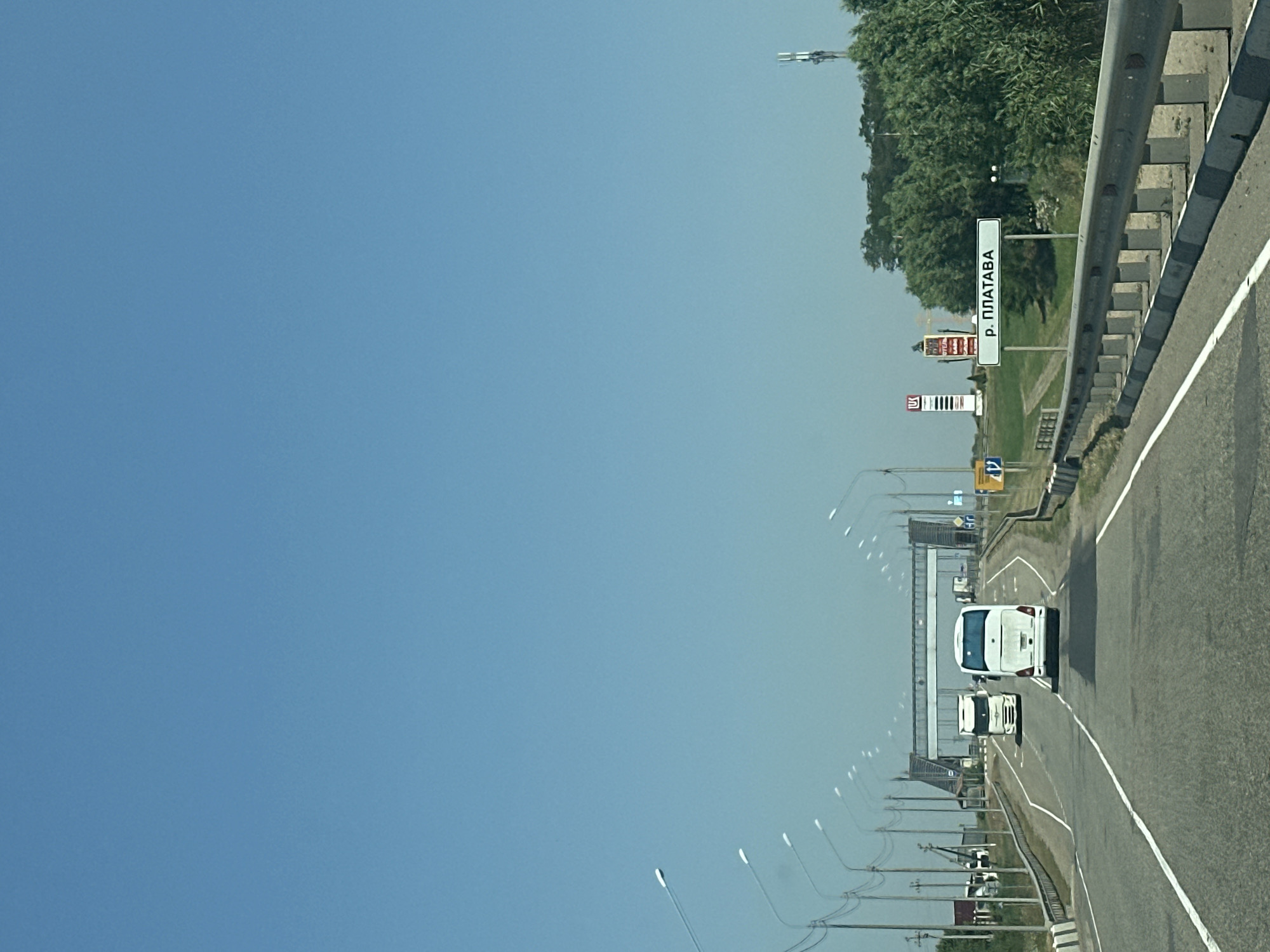 The image shows a rural road in Yugo-Severnaya, Russia. The road is asphalt and has a white line painted down the center. There are two vehicles on the road: a white bus and a white truck. The bus is in the foreground and is facing away from the camera. The truck is behind the bus and is also facing away from the camera. There are trees and a communication tower on the right side of the road. A sign with Russian text is visible on the right side of the road, indicating "р. ПЛАТАВА" (r. PLATAVA). There are streetlights lining the road on both sides. In the distance, there is a large pedestrian bridge crossing the road. The sky is clear and blue. The overall impression is one of a sunny day on a highway in a rural area.