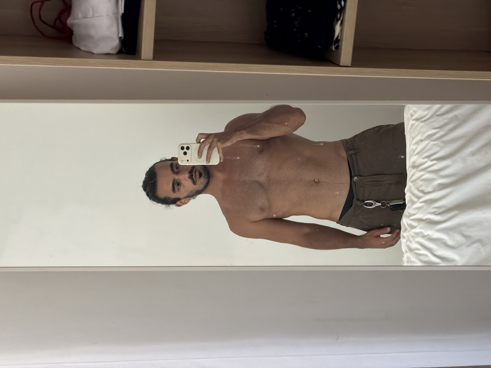 A man is taking a selfie in a mirror. The mirror reflects his torso, face, and phone. He is wearing pants, and he appears to be indoors, perhaps in a bedroom or closet area, given the presence of shelving above the mirror. The location is Kuta Selatan, Indonesia.