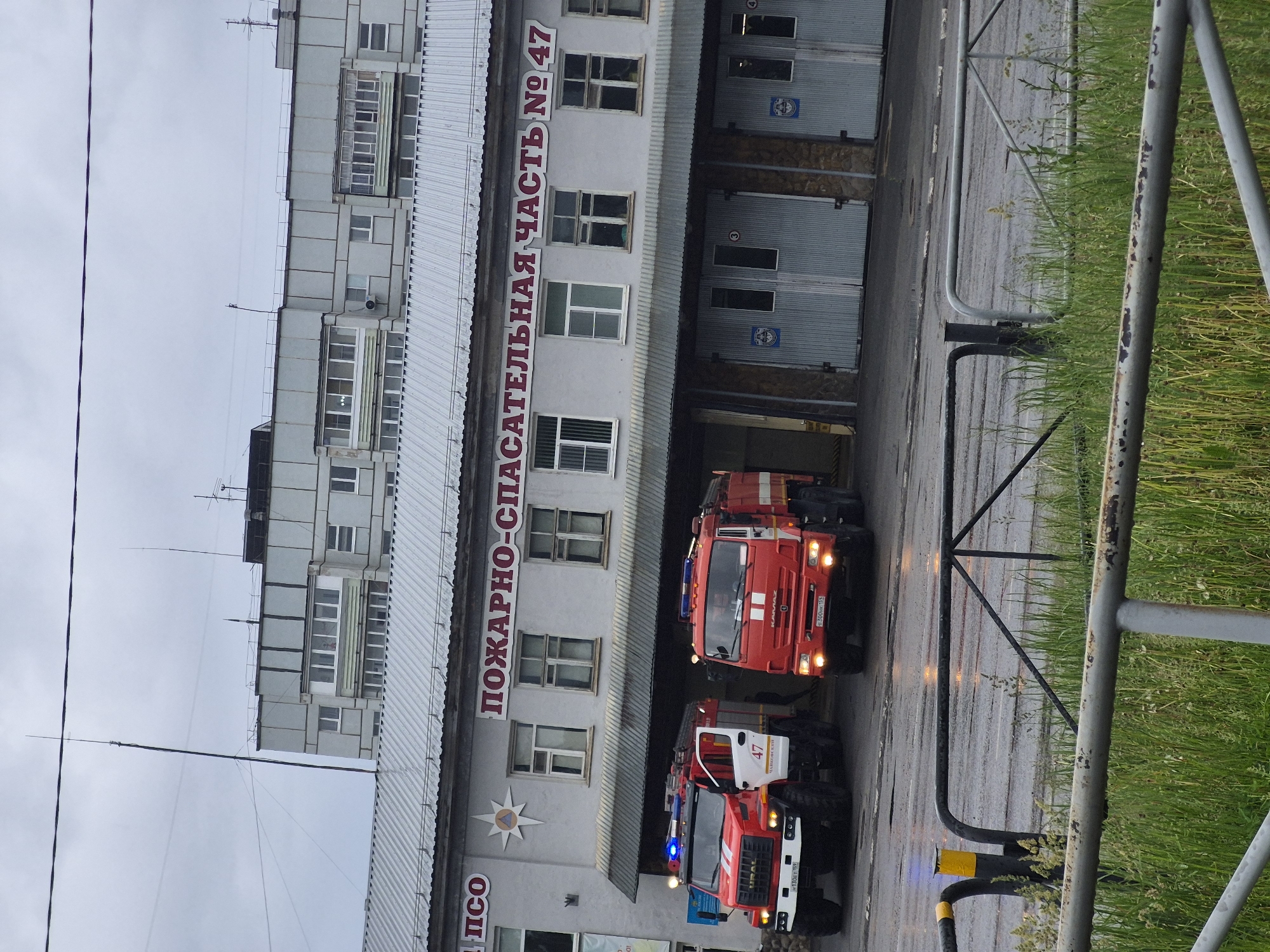 The image captures an exterior view of a fire and rescue station, likely located in Chaikovsky, Russia. Two red fire trucks are prominently featured on a wet asphalt driveway in front of the station's garage bays.
The station building is a multi-story structure with a light-colored facade. Above its garage doors, a large red sign in Cyrillic reads "ПОЖАРНО-СПАСАТЕЛЬНАЯ ЧАСТЬ № 47," which translates to "Fire-Rescue Unit No. 47." To the left of this sign, an emblem resembling a star with a compass-like design is visible, along with the partially obscured text "ПСО" (likely referring to the Emergency Rescue Service).
The fire truck on the left is a white and red Ural model, bearing the number "47" on its side and displaying an illuminated blue flashing light. Its license plate is "М 130 ЕТ 59". The second truck, positioned slightly behind and to the right, is a red Kamaz model, also with its lights illuminated, and its license plate is "Е 200 НМ 59". The trucks appear to be either preparing for deployment or have recently returned to the station.
The scene is set on an overcast day, with a grey, cloudy sky. The wet appearance of the asphalt suggests recent rain. To the right of the driveway, a metal fence separates the operational area from a strip of green grass. No people are visible in the image. The overall impression is of an active rescue service facility under potentially damp weather conditions.