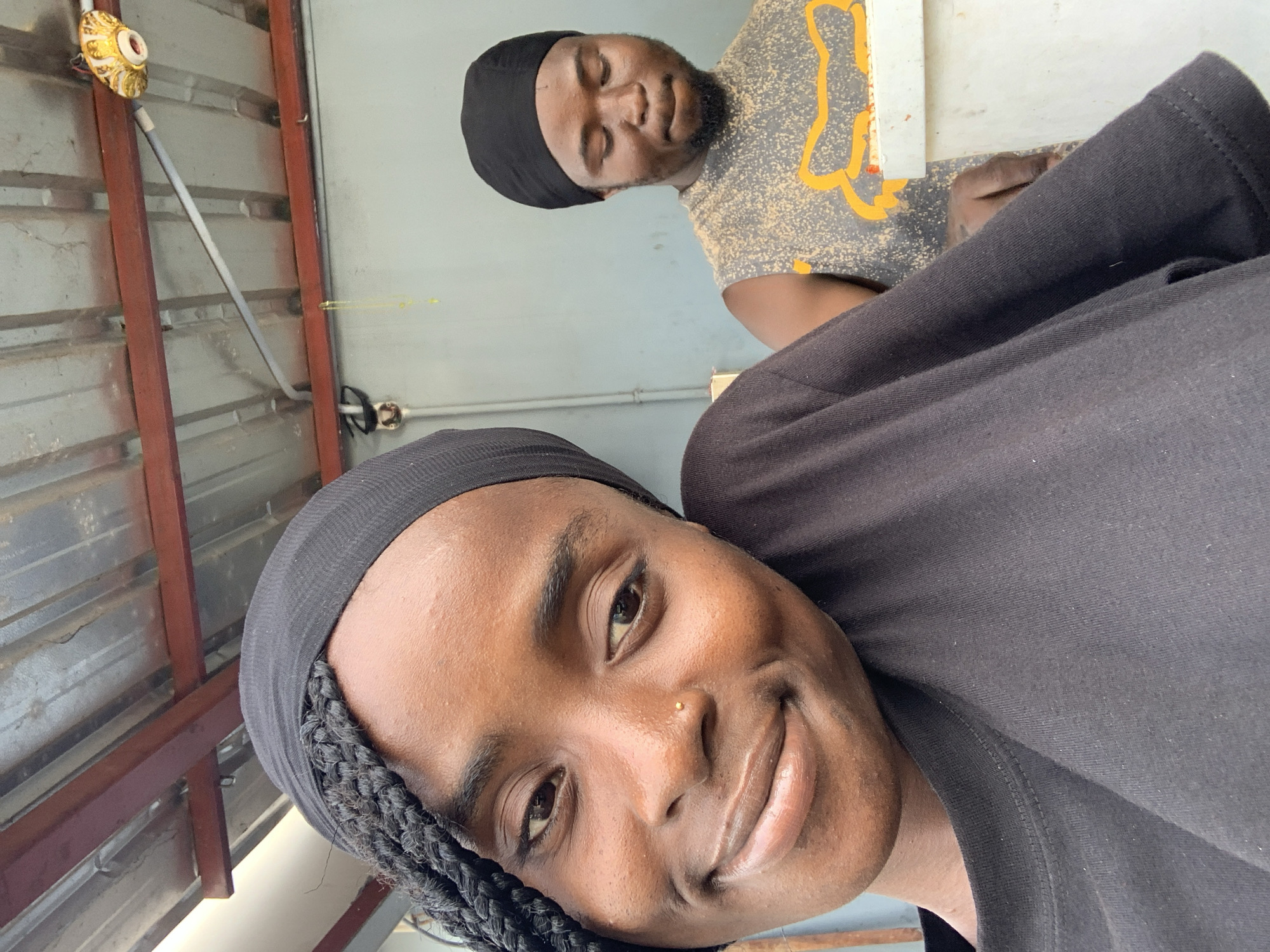 This image, taken in Adankolo, Nigeria, features two individuals in what appears to be an indoor or covered setting. The primary subject, a person with dark skin, is in the foreground, looking directly at the camera with a bright smile. This individual wears a black head covering, revealing dark braided hair on the side of the face, and a black t-shirt. A small golden nose stud is visible on their right nostril.
Behind and slightly to the side of the foreground subject, another person with dark skin is visible, wearing a black head covering and a light-colored t-shirt with a speckled yellow and orange pattern. This individual has a beard and appears to have their eyes closed or cast downwards, showing a calm and relaxed expression.
The background features a corrugated gray wall or ceiling structure with reddish-brown metal support beams. A decorative light fixture or utility box with gold and white ornate details is mounted on the wall in the upper left section of the frame, connected by a gray conduit. Behind the second person, a plain light blue-gray wall is visible. Exposed electrical wiring and an outlet are also present on the walls. The lighting is bright and even, suggesting it is daytime. There is no visible text in the image.