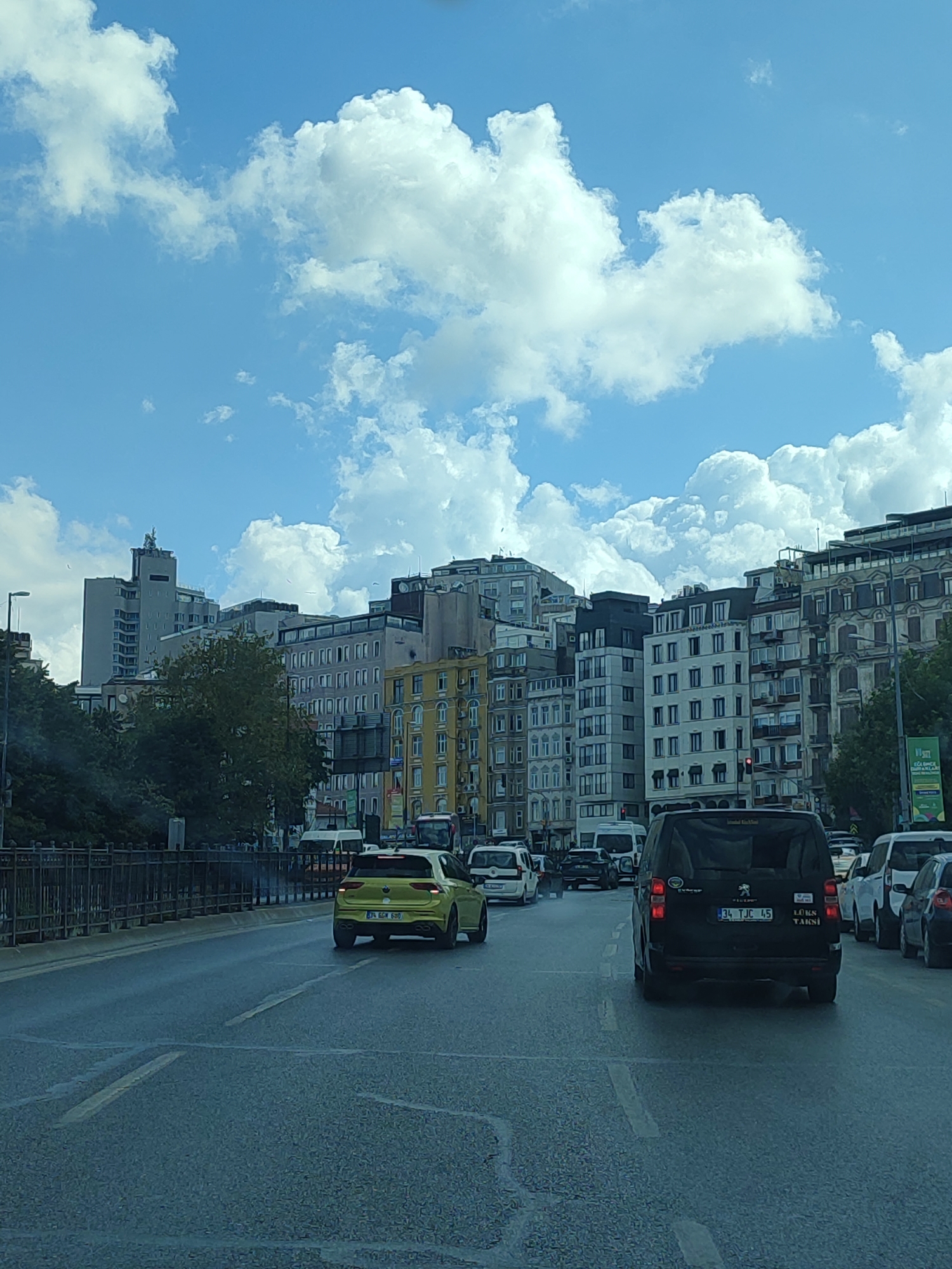 The image captures a view from a vehicle traveling on a street in the Beyoğlu district of Istanbul, Turkey, with a backdrop of city buildings and a bright, cloudy sky. In the foreground, the asphalt road is wet, possibly from recent rain, with visible lane markings. Several vehicles are present: a yellow car in the left lane, a black van in the right lane with "LÜKS TAKSİ" visible on its rear, and other cars and vans further ahead and to the right. The middle ground features a stone bridge with metal railings on the left, and on the right, a row of multi-story buildings. These buildings exhibit a variety of architectural styles, from modern high-rises to older structures with ornate facades and balconies, painted in neutral tones, yellow, and cream. Trees with green foliage are interspersed between the buildings. The sky above is a vibrant blue, dotted with large, white cumulus clouds. A traffic light is visible on one of the buildings.