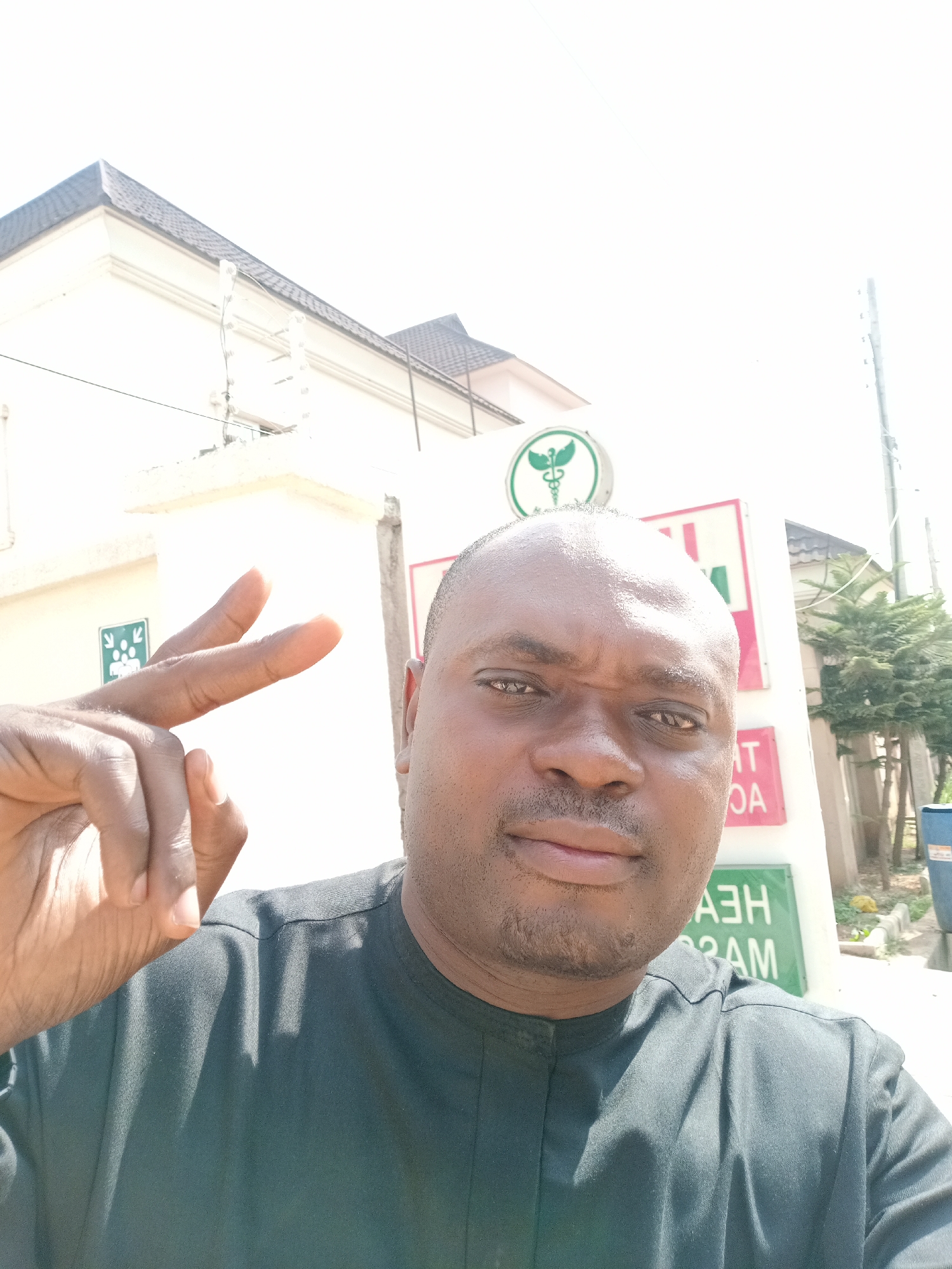 This bright, sunlit selfie captures a dark-skinned man outdoors in Gwarinpa, Nigeria. The man, who has a shaved head and short beard, occupies the center-right of the frame, looking directly at the camera with a neutral to slightly serious expression. He is wearing a dark, possibly black or dark green, collared shirt. His left hand is raised to the left of his head, forming a "peace" or "victory" sign with his index and middle fingers extended.
The background features architectural elements and signage, indicative of a public or commercial setting. A prominent white building with a dark grey tiled roof is visible, along with parts of two distinct signs. A circular green sign in the upper center displays a white Caduceus symbol, commonly associated with medicine or pharmacies. Below and to the right of this, a larger red and white sign shows mirrored text which, when reversed, appears to read "HEALTH" (partially visible as "HEA") and "MASS" (partially visible as "MAS"), suggesting "HEALTH MASS" or a similar phrase. Another small green sign to the left of the man's raised hand shows white stick figures with arrows, possibly indicating an assembly point or direction. To the far right, a green shrub or small tree, a blue container, and a utility pole are visible, confirming an outdoor location during what appears to be a bright, clear day.