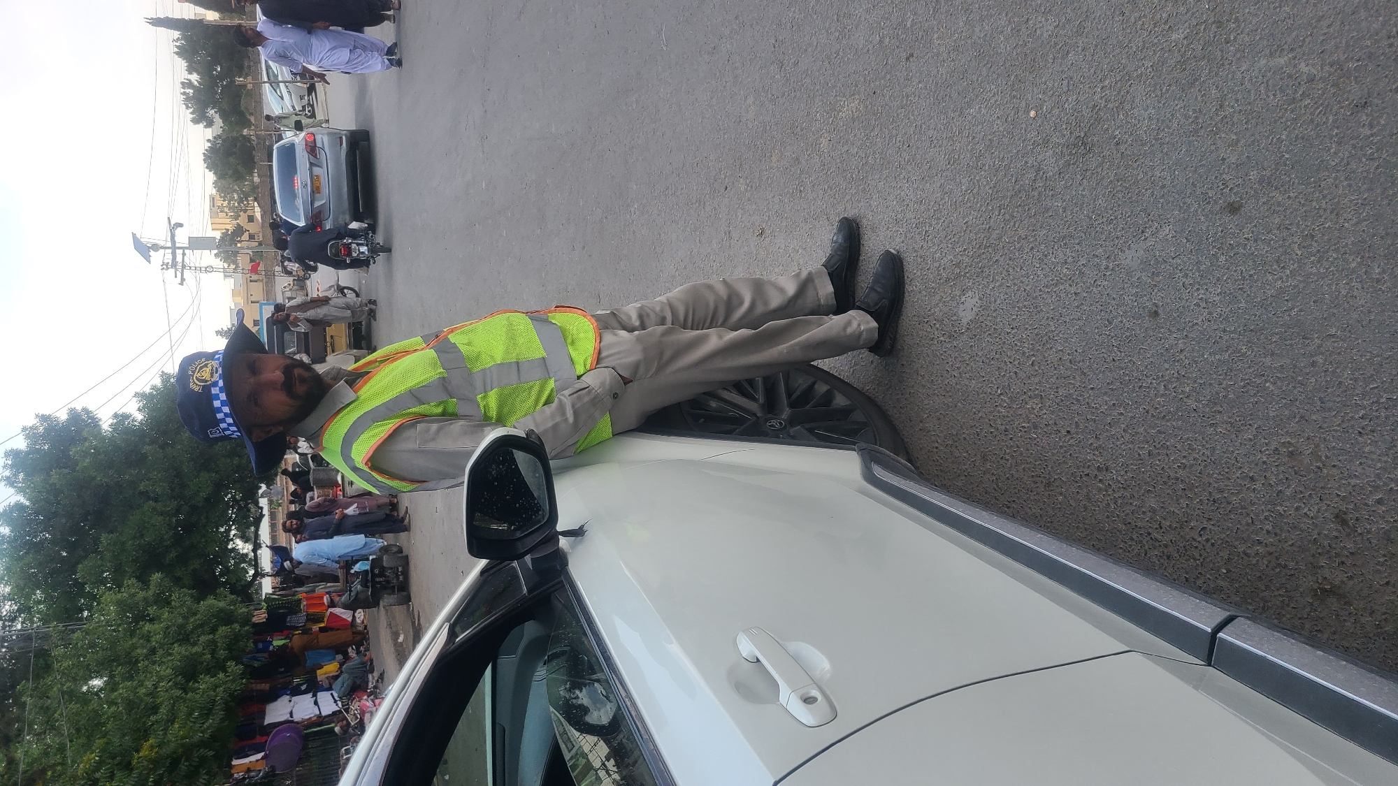 A traffic police officer in Quetta City Tehsil, Pakistan, lies on the hood of a white car with his legs resting on a tire. He is wearing a blue and white checkerboard hat, a yellow and green reflective vest, and tan pants. The background shows a street scene with pedestrians, vehicles, trees, and buildings.
