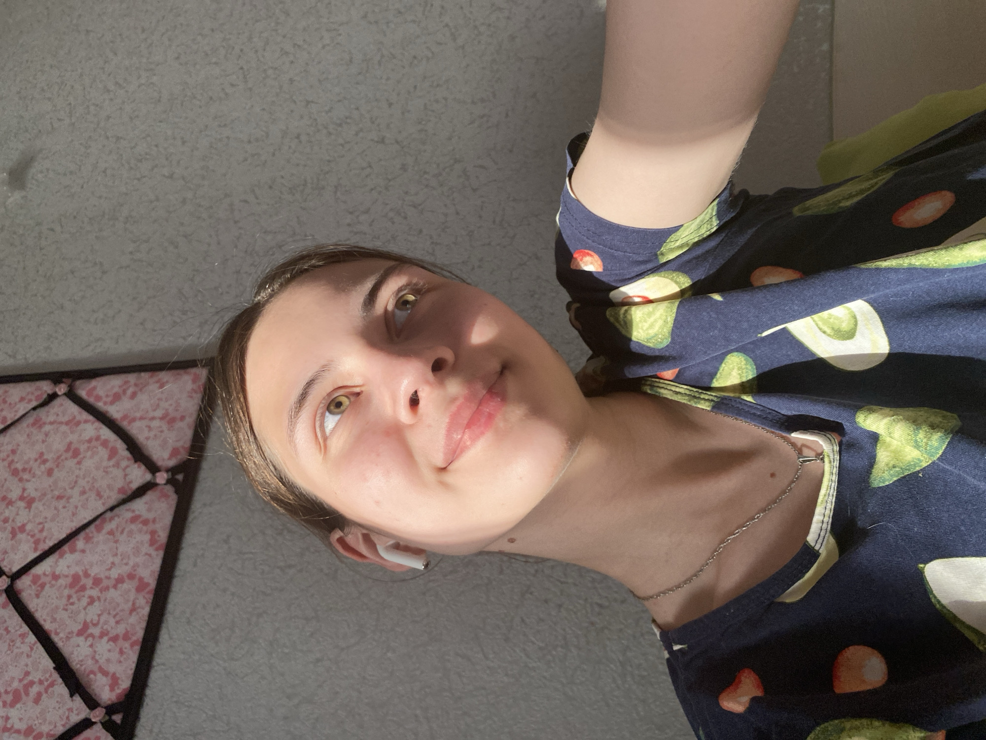 A person is viewed from a low angle, looking up at the camera. The person has light skin, light brown hair pulled back, and green eyes. They are wearing a dark blue shirt with a pattern of avocados and tomatoes. A silver chain necklace is visible around their neck, and a white wireless earbud is in their left ear. Part of a wall is visible in the background, with what appears to be a patterned fabric or headboard in the lower left corner. The lighting appears to be natural sunlight. The location is not possible to determine from this image.