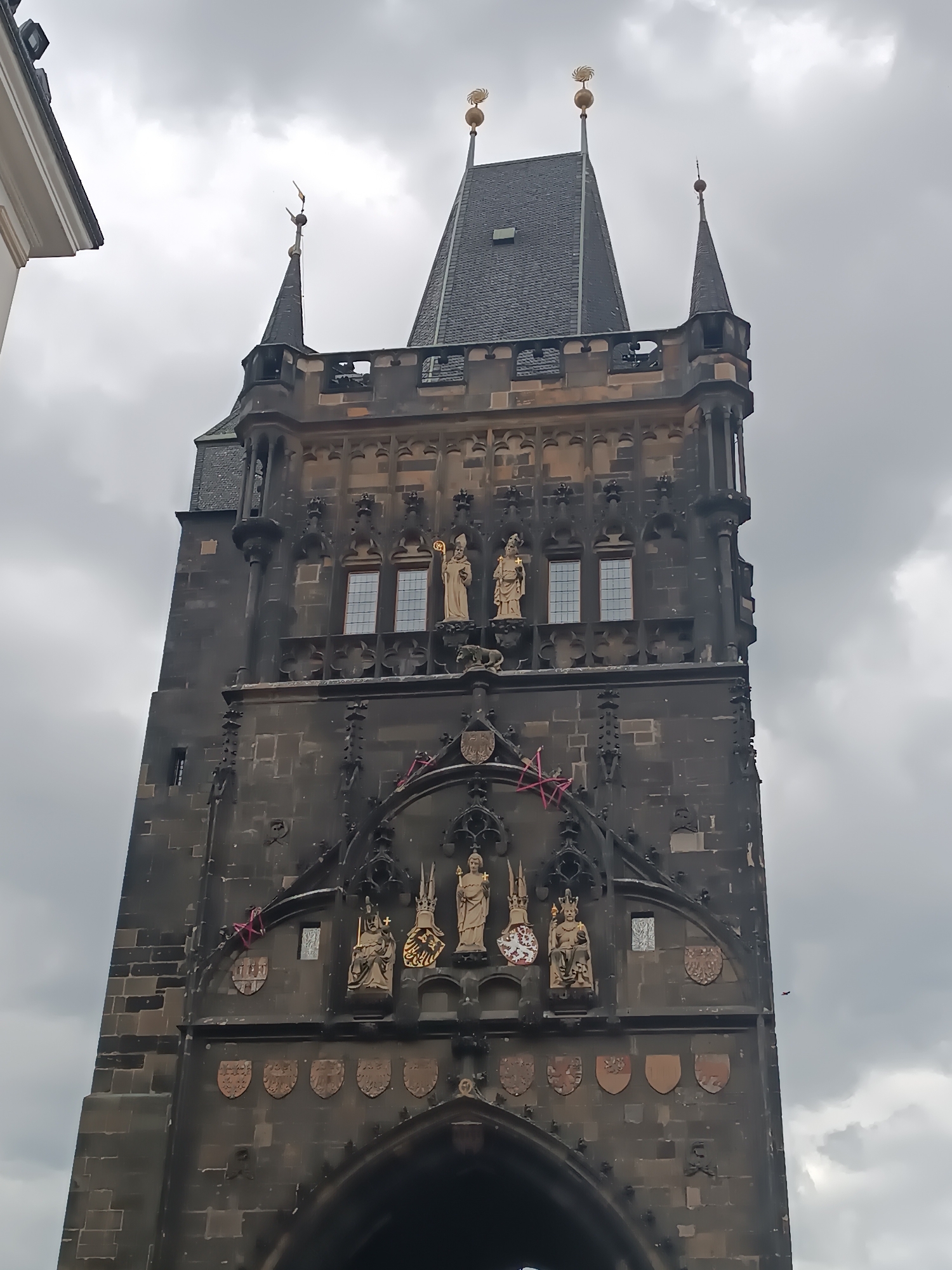 The image shows a detailed view of the Old Town Bridge Tower in Prague, Czechia, a Gothic stone structure. The tower is ornately decorated with sculptures of saints and coats of arms. Above the main archway, several statues are visible, including one depicting a crowned figure holding a scepter and orb, and another of a saint with a halo. Above these, further statues are positioned in niches, some with gold accents. The tower has a steep, dark gray tiled roof with spires topped with golden decorative elements. The sky is overcast with gray clouds. The bottom portion of the tower, featuring a large archway and more decorative shields, is also visible.
