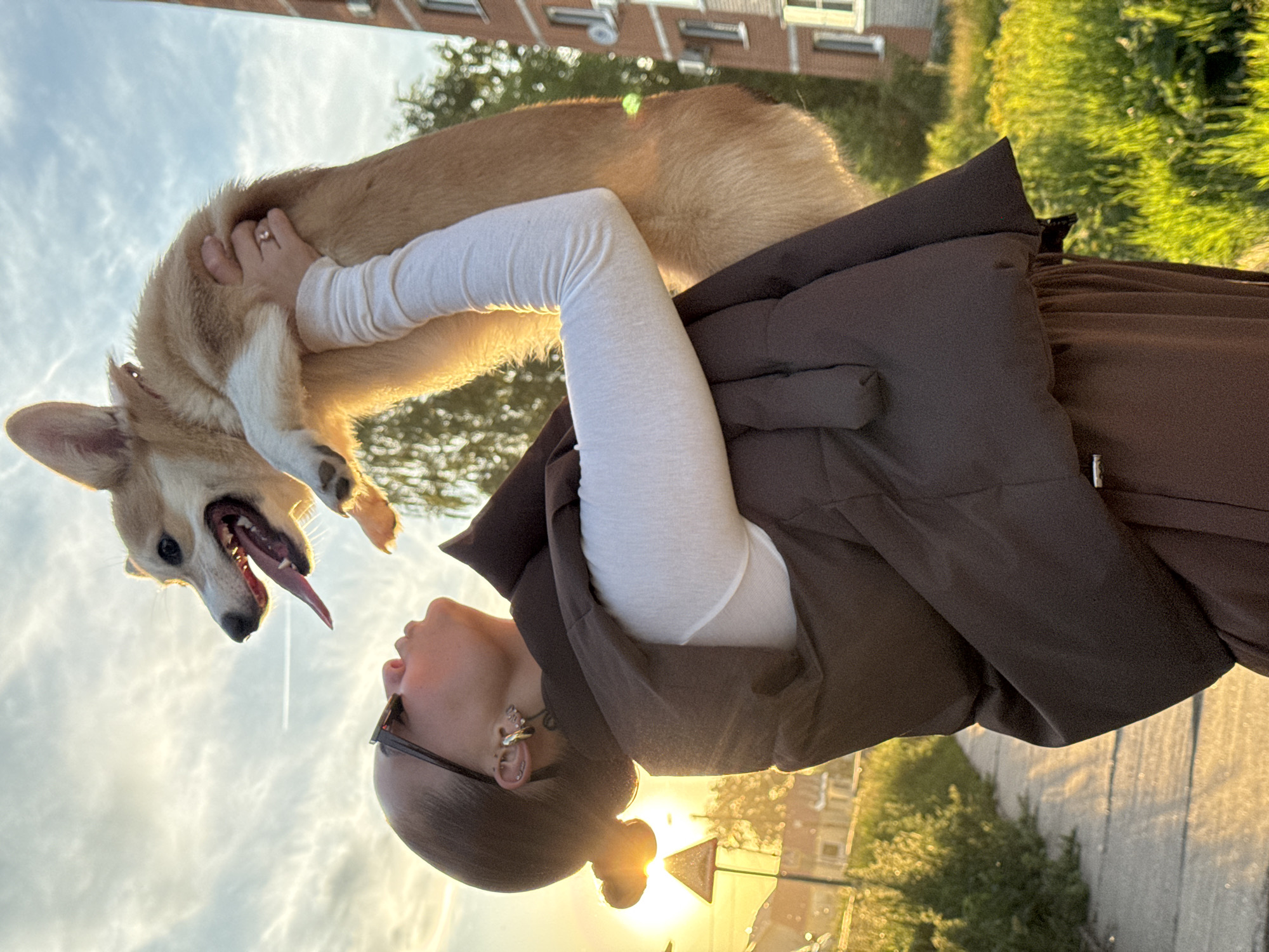 An eye-level, portrait-oriented photograph captures a person holding a Pembroke Welsh Corgi dog against a warm, backlit sky at sunset or golden hour in Chaikovsky, Russia.
The central subjects are a person and a light brown and white Corgi. The person, positioned on the right, looks up at the dog. They have their hair pulled back in a bun and wear black-framed glasses, a distinctive silver and black earring on their left ear, and a ring on their left hand, which is visible petting the dog's back. They are dressed in a dark brown sleeveless jacket or vest over a white long-sleeved top. The Corgi is held aloft on the left, looking down at the person with an open mouth, extended tongue, and perked ears, conveying excitement and playfulness. The dog's fur glows from the backlight.
The scene is outdoors, likely in an urban or suburban setting. Out-of-focus buildings are visible in the upper background, with green foliage and trees below them. The ground visible in the lower right is a paved surface, possibly asphalt or concrete. The time of day is clearly sunset or golden hour, with a low sun casting a warm, bright glow across the sky and silhouetting parts of the subjects. The sky features scattered clouds, indicative of fair weather. The activity depicted is an affectionate interaction between the person and their dog. The overall emotion conveyed is one of joy and connection.
No visible text is present in the image.