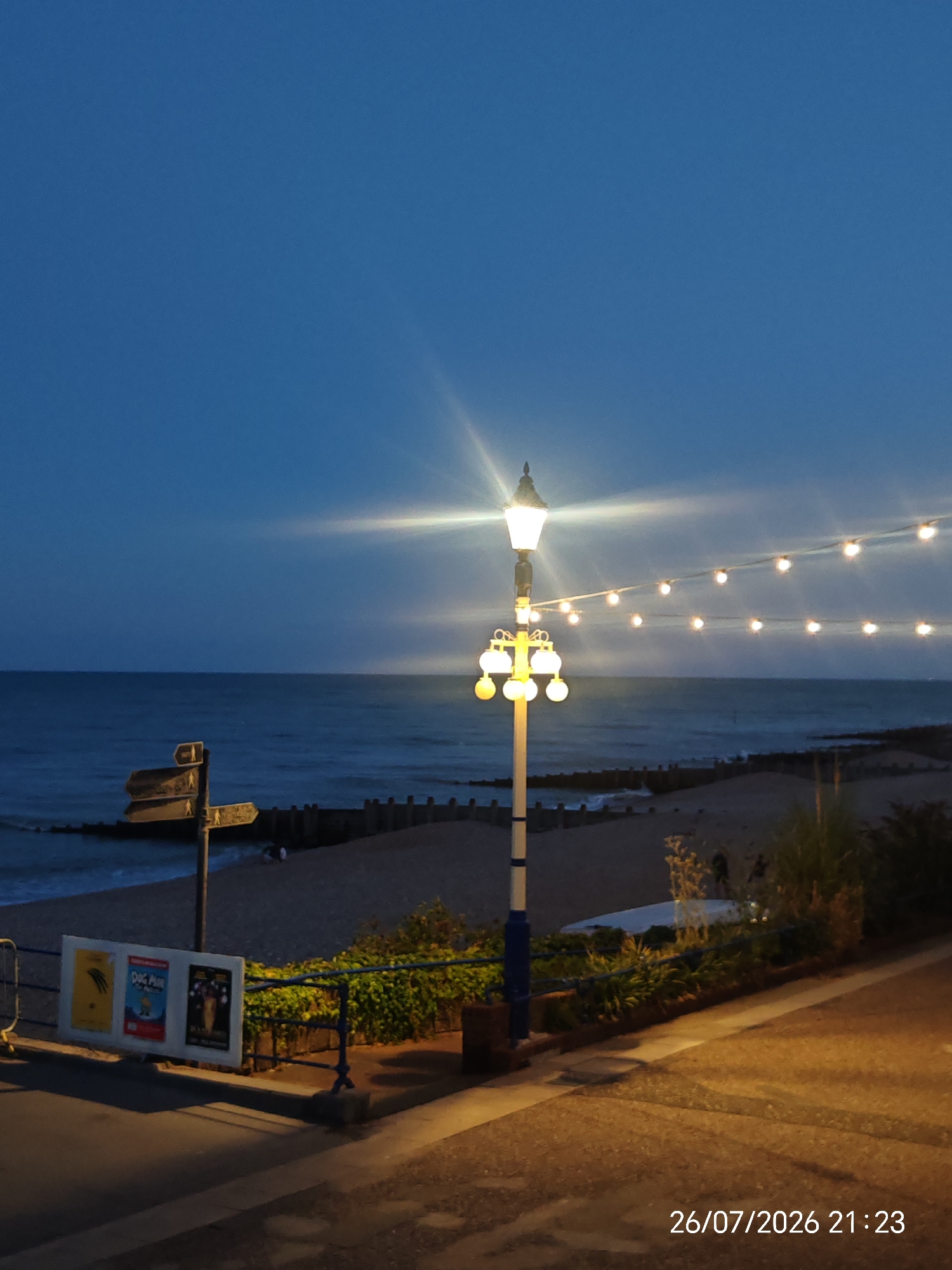 The image captures a dusk scene at an Eastbourne beach, featuring a sandy and pebbly shoreline bordering a calm sea under a dark blue sky. A streetlamp with multiple illuminated globes stands prominently in the foreground, adorned with a string of fairy lights extending across the frame. To the left, a signpost with directional arrows indicates paths, and three framed posters, one labeled "Dog Man," are mounted on a railing. The pathway in the immediate foreground is paved and illuminated by the warm glow of the streetlights, reflecting on the wet surface. The overall atmosphere is tranquil, with a few indistinct figures visible on the beach in the distance. A date and time stamp, "26/07/2026 21:23," is visible in the bottom right corner.
