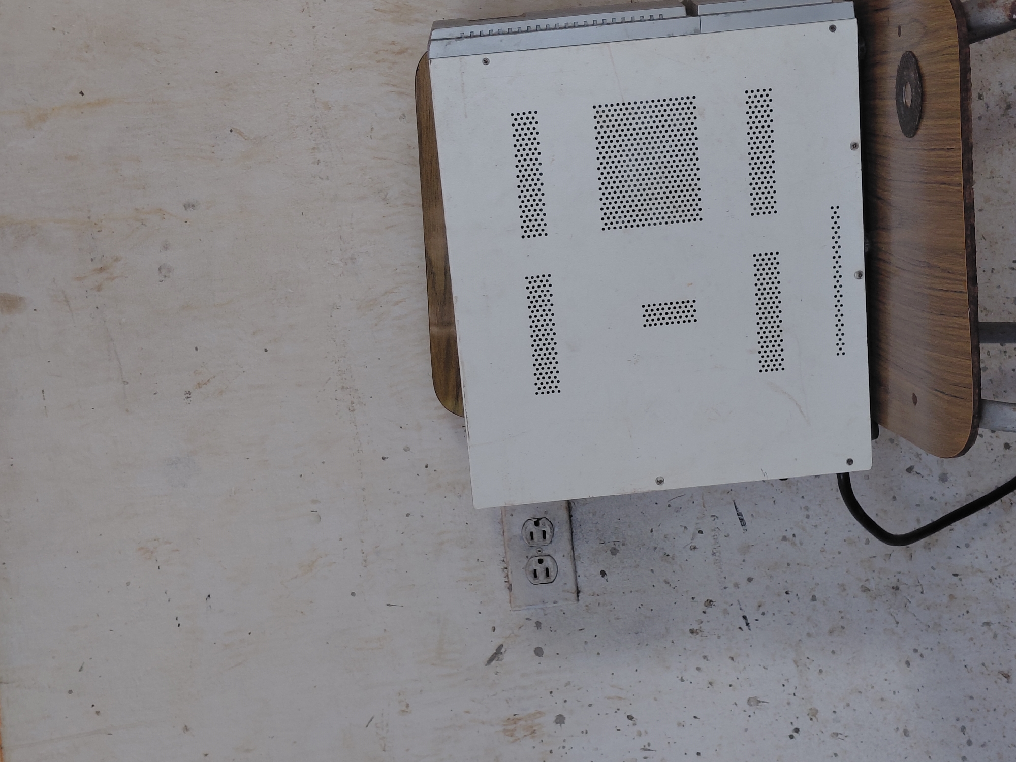 The image depicts a close-up, eye-level view of an electrical outlet and a white electronic device with a perforated casing, positioned on a wooden surface. The wall behind the outlet and device is dirty and stained, suggesting an unmaintained environment. The outlet appears to be a standard dual-receptacle type. The electronic device, possibly a computer tower or a similar piece of equipment, has a light-colored, perforated metal exterior, with various patterns of circular holes for ventilation. A black power cord is visible extending from the device. A small, dark, circular object, possibly a washer or a small disc, rests on the wooden surface near the electronic device. The background wall is a pale, off-white color, marked with numerous dark spots, smudges, and faint streaks, indicating wear and tear. The lighting in the scene is neutral, with no strong shadows. The image does not contain any people or direct indicators of the specific location within Santa Ana, Venezuela, beyond the assumption provided in the prompt.