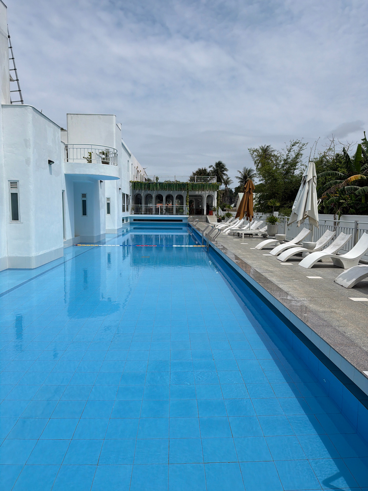 A long, rectangular swimming pool with blue tiled bottom occupies the foreground of the image. The pool is sectioned by lane ropes. To the right of the pool is a grey tiled deck with white lounge chairs and closed white and brown umbrellas. In the background, a white building with a balcony and a ladder on its side runs along the left of the pool. To the right of the building, a covered walkway with arched openings is visible, with palm trees and other greenery behind it. The sky is cloudy. The image appears to be taken at a resort or hotel on the island of Phú Quốc, Vietnam, given the architectural style and tropical vegetation.