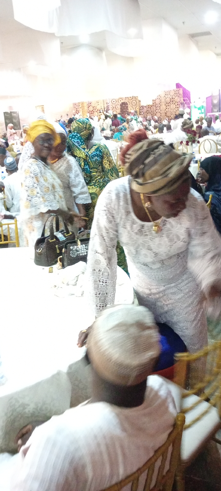 The image depicts an indoor event, likely a gathering or celebration, taking place in Gandu Albasa, Nigeria. The scene is filled with people dressed in traditional attire, predominantly women wearing vibrant headscarves and elaborate dresses. Several individuals are seen standing, while others are seated at tables covered in white cloths. The background features a decorated wall with intricate patterns and subtle lighting, suggesting a formal or festive occasion. In the foreground, two women are the central focus; one wears a yellow headscarf and a white lace outfit, while the other is adorned in a brown and white striped headscarf and a white lace dress. Bags are placed on a table in front of them. The overall atmosphere appears to be social and lively.