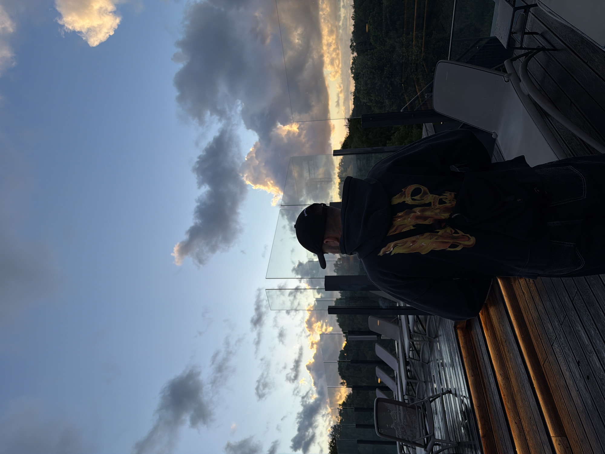 The image depicts a man wearing a black baseball cap and a black hoodie with a flame graphic on the back, standing on a wooden deck with illuminated stairs. He is facing away from the camera, looking towards a cloudy sky during what appears to be sunset. In the background, there are glass railings, lounge chairs, and lush green trees. The scene suggests an outdoor relaxation area, possibly a resort or hotel in Kołobrzeg, Poland, given the overcast sky and the presence of lounge chairs often found near bodies of water. The lighting indicates a transition from day to evening, with the clouds catching the warm hues of the setting sun. The deck is wet, possibly from recent rain, adding to the atmosphere.