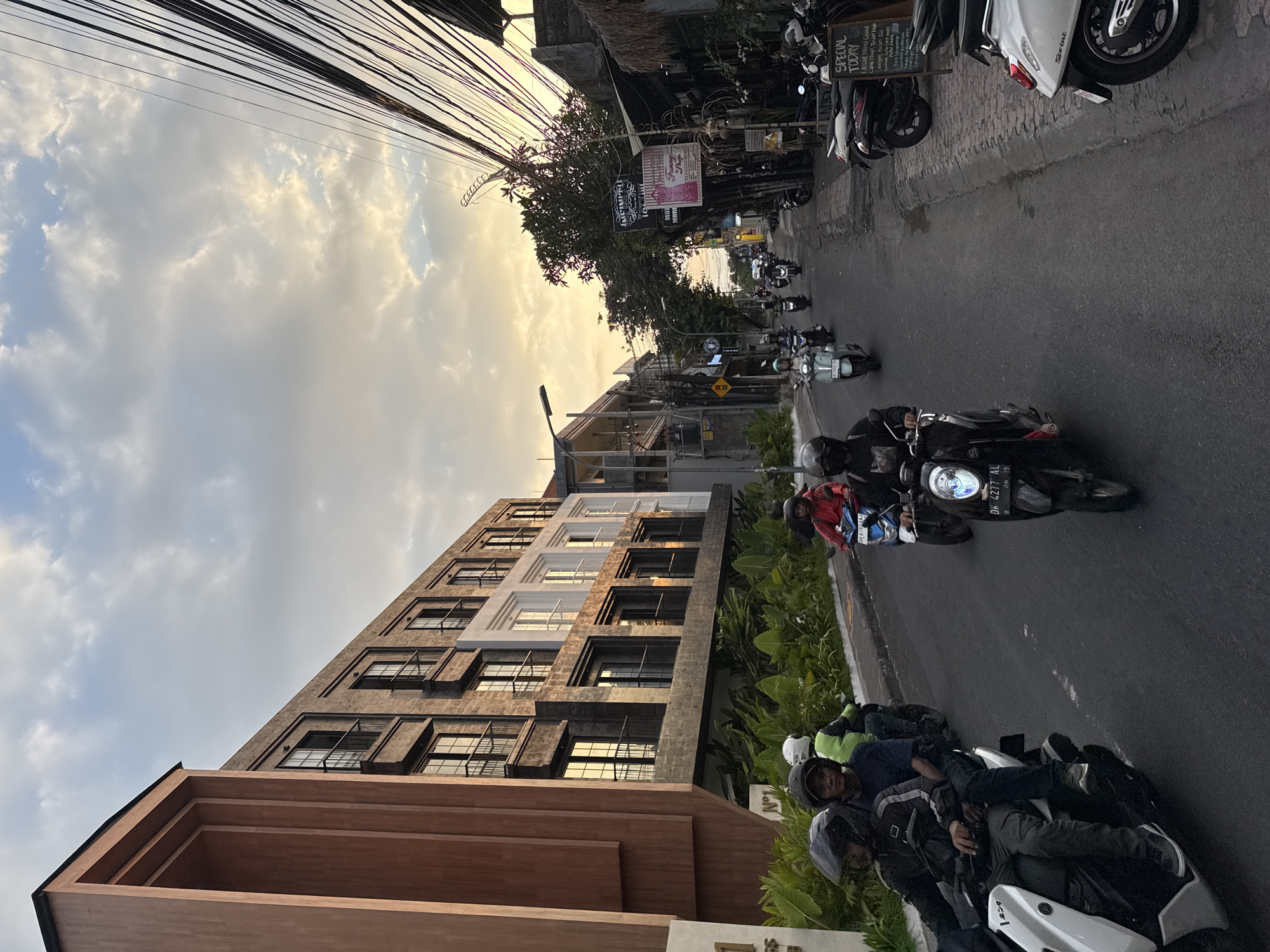 The image depicts a street scene in Kuta, Indonesia, during what appears to be sunset. A multi-story building with modern architecture, featuring wooden and stone elements and numerous windows, dominates the left side of the frame. The building has a pronounced cantilevered section extending over a sidewalk lined with lush green plants.
The street is populated by several people riding motorbikes, a common mode of transportation in the area. The riders are wearing helmets, and some are carrying passengers. The motorbikes are of various models, including scooters and more traditional styles.
On the right side of the street, commercial establishments are visible, with signs and awnings. One sign, in particular, advertises "SPECIAL TODAY" with a handwritten menu, suggesting a food establishment. Overhead, a dense network of electrical wires crisscrosses the sky, a characteristic feature of many urban landscapes in Southeast Asia.
The sky displays a gradient of colors, from pale blue to warm oranges and yellows, indicating the setting sun. The overall atmosphere is that of a busy, active street at the end of the day, with a blend of modern development and the vibrant local life of Kuta.