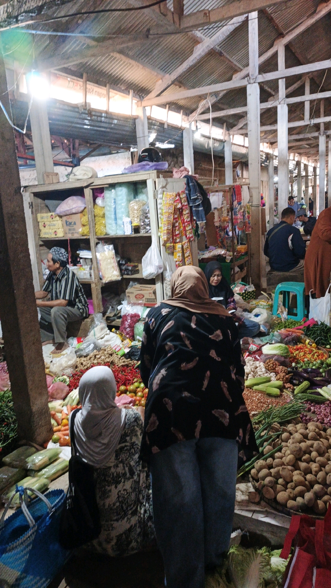 The image depicts a bustling market scene in Cibenda, Indonesia, under a corrugated metal roof supported by wooden and concrete pillars. The market is filled with an assortment of fresh produce, including potatoes, red chilies, tomatoes, onions, and various vegetables, displayed on the ground and on wooden stalls. In the foreground, two women wearing hijabs are partially visible, one in a patterned dress and the other in a black patterned top. A man in a striped shirt and a patterned skullcap sits on a stool, possibly a vendor, to the left, with shelves of packaged goods behind him. Several other individuals are seen in the background, engaged in shopping or tending to their stalls. Bags and baskets are scattered around, suggesting active commerce. The overall atmosphere is one of a lively local marketplace.