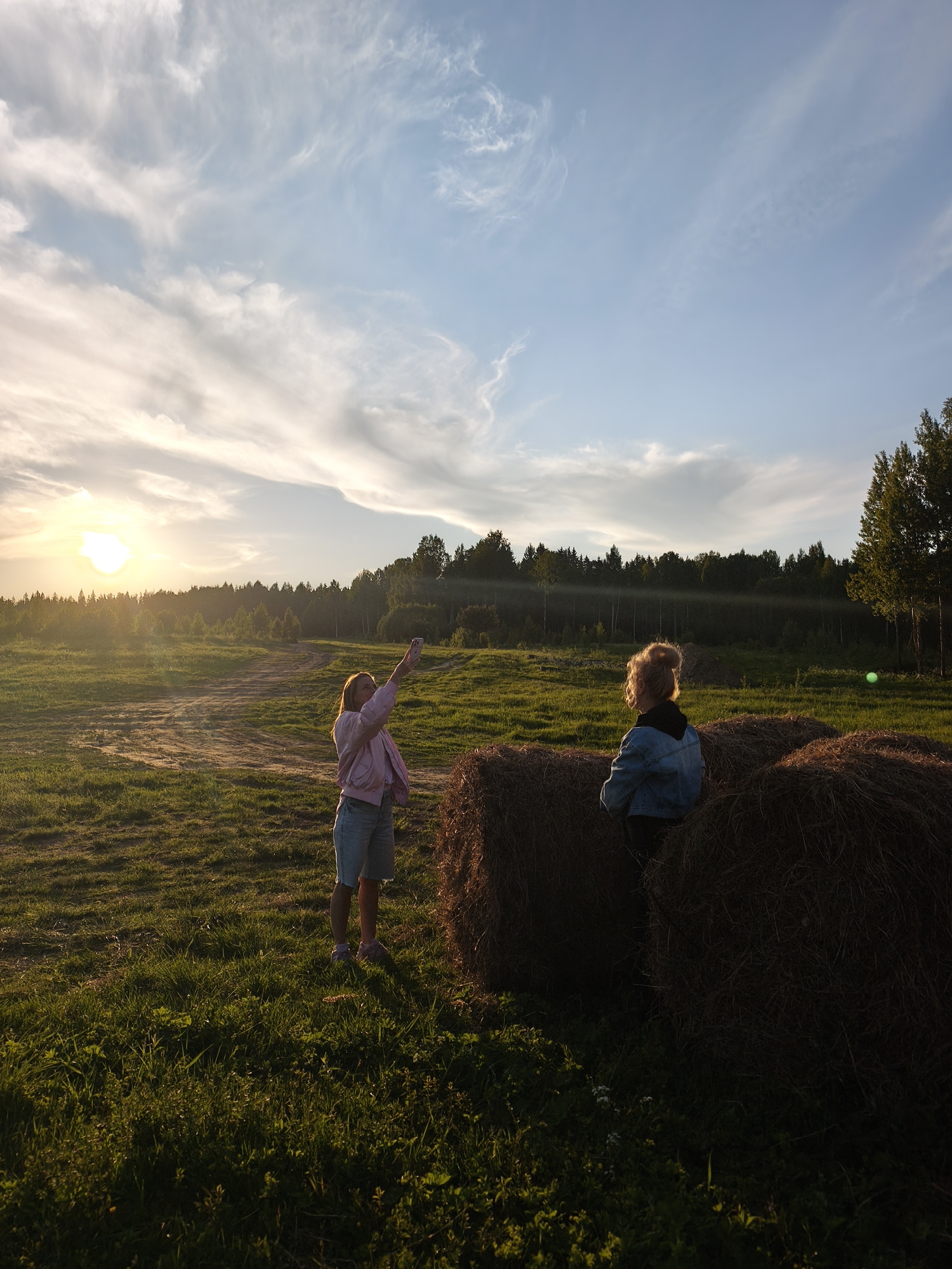 The image captures a serene, golden hour scene in a rural landscape near Стеклянный, Russia. The setting sun, a brilliant orb on the left horizon, bathes the field in a warm, low light, casting long shadows and creating a sense of peace.
Two young individuals are present in the foreground. On the left, a person with long hair, wearing a light pink jacket, light blue shorts, and pink sneakers, stands holding a smartphone aloft, seemingly capturing the sunset or taking a selfie. Her posture suggests active engagement with the moment. To the right, another person with curly, reddish-blonde hair tied in a bun, dressed in a blue denim jacket over a dark top, is seated on or leaning against a large, round hay bale. This individual is turned slightly away, looking towards the right side of the frame, possibly observing the surroundings or the other person. Several large hay bales are stacked in the right foreground, providing texture and a rustic element to the scene.
The ground is covered in lush green grass, with visible dirt paths winding through the field. In the mid-ground, a dense line of dark green forest stretches across the horizon, silhouetted against the bright sky where the sun is setting. Above, the sky transitions from bright, warm hues near the sun to a soft blue, adorned with wispy, scattered clouds. The overall atmosphere is tranquil and relaxed, with the natural beauty of the countryside emphasized by the dramatic lighting of the sunset. There is no visible text in the image.