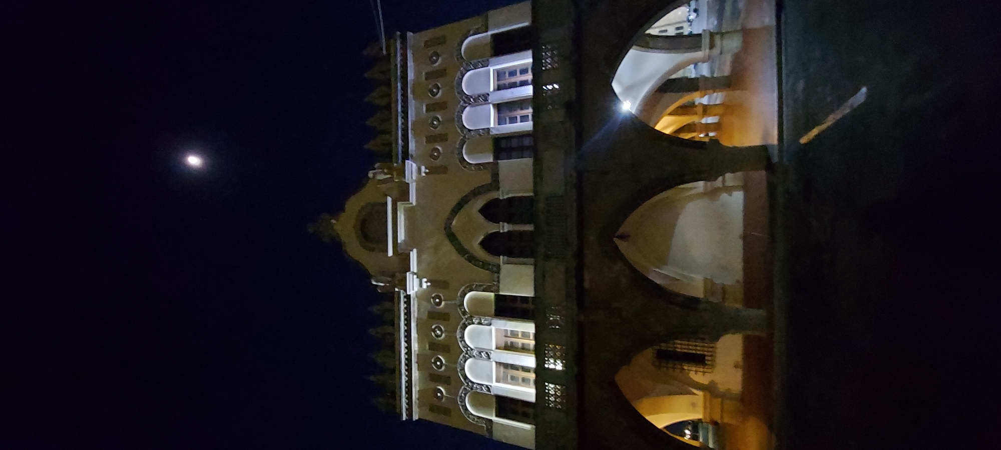 A low-angle, full-frame, vertical, nighttime exterior shot captures the illuminated facade of the Municipal Market of Rhodes, Greece. The building, constructed of stone and painted a light tan color, features a prominent central section with decorative arched windows and a crenellated roofline. Below this, a series of arches creates a covered walkway. The sky above is a deep, dark blue, with a small, bright full moon visible towards the top center. The ground in front of the market is dark and appears wet, reflecting some of the light from the building.