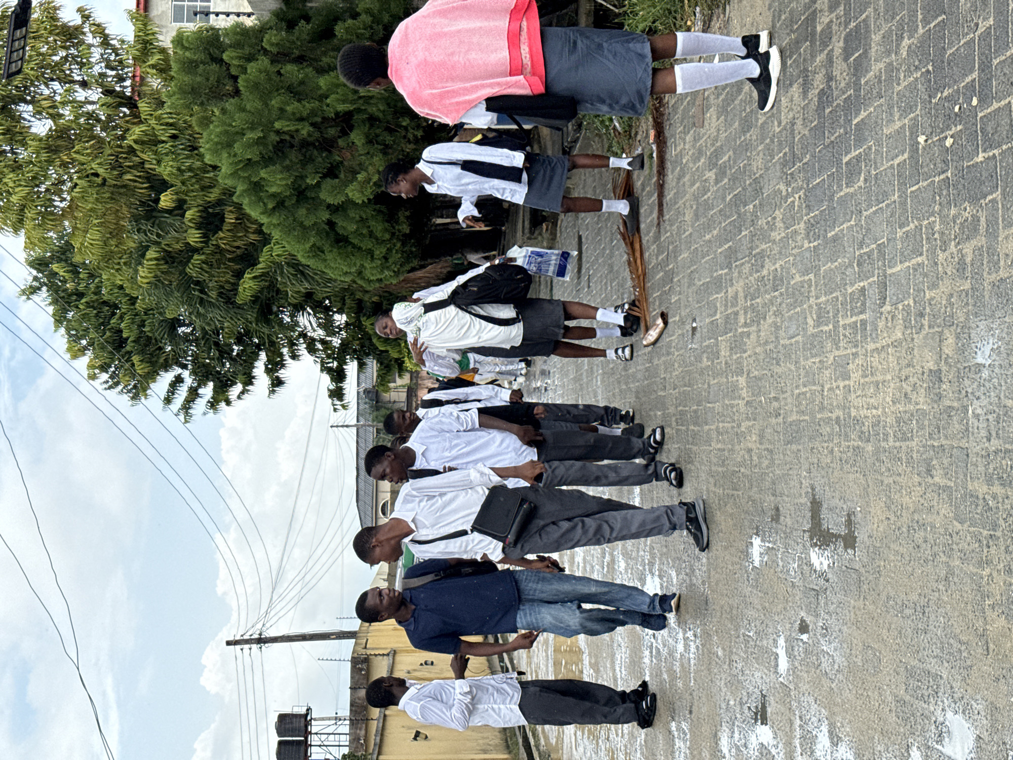 A horizontal shot depicts a line of people standing on a brick road in Ado, Nigeria. At the front, a man in a blue shirt and jeans stands. Behind him are men wearing white shirts and trousers. Further back, three students wearing white shirts, gray shorts, and knee-high white socks stand in a row. A tree is to the left of the students. Overhead power lines and a water tank are visible in the background. The sky is cloudy.