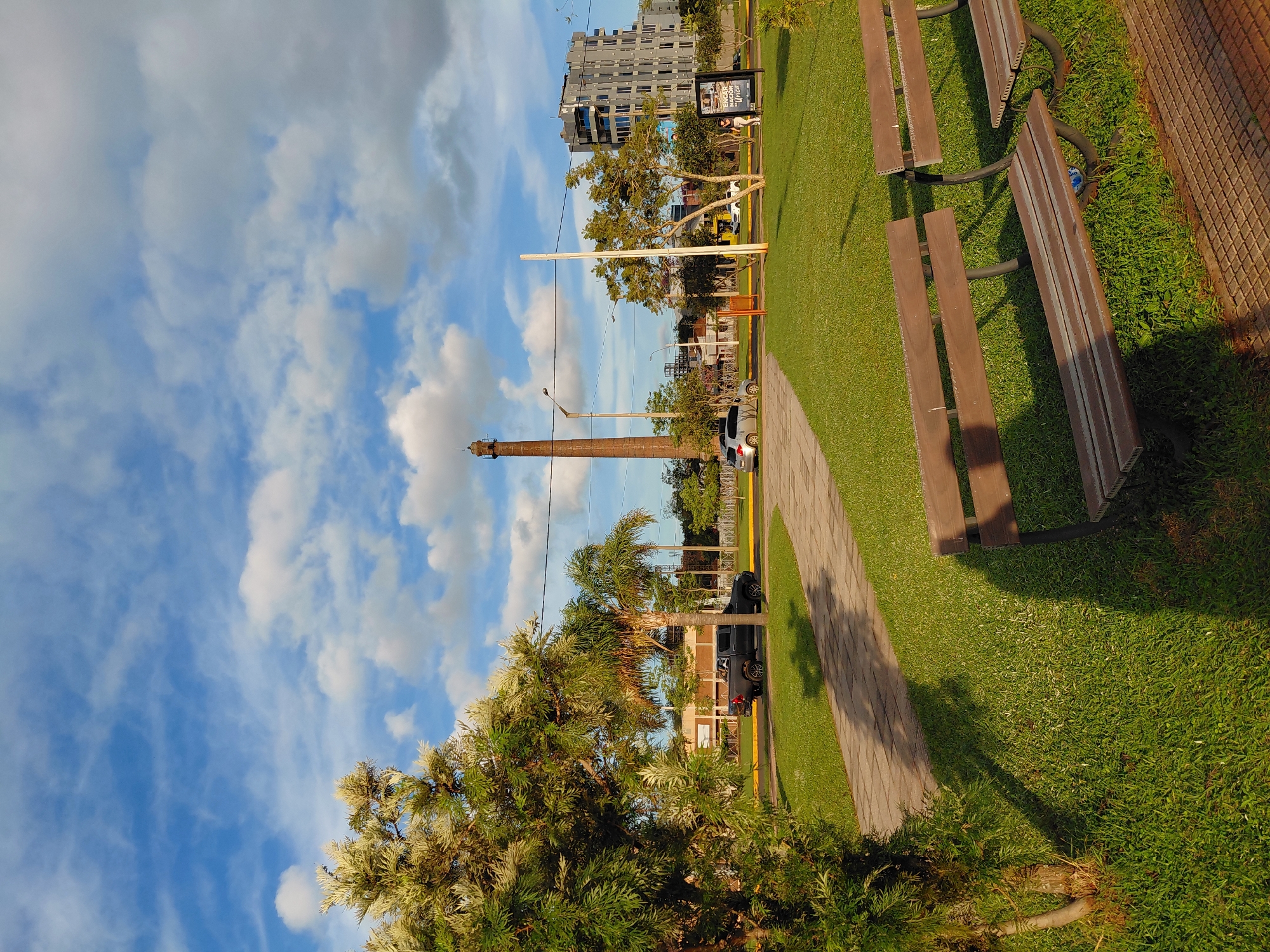 This is an outdoor, eye-level, full shot of a park area in Encarnacion, Paraguay, during the daytime. The foreground features two empty, wooden park benches on a grassy area. A paved walkway with a brick border is visible to the left and in front of the benches. The middle ground shows more green grass and trees, including a large evergreen tree on the left and palm trees behind it. A dark-colored pickup truck is parked on the right side of a paved road that runs horizontally across the image. Further in the background, a tall, reddish-brown brick smokestack stands prominently. To the right of the smokestack, a modern, multi-story grey building with many windows is visible. A few other vehicles and a person are also present on the road and sidewalk in the distance. The sky is blue with scattered white and grey clouds. The overall lighting suggests it is late afternoon, with long shadows cast on the grass.
