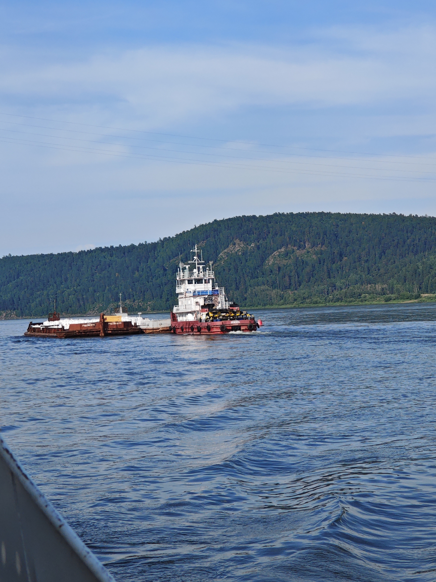 In the city of Pristan', Russia, a vessel is navigating a wide body of water. The vessel, likely a tugboat, is red and white, with a superstructure that appears to be several stories high. It is pushing a barge that is loaded with what looks like ice or some sort of white material. The barge itself is brown and metal. The water is choppy with visible ripples. In the background, a densely forested hillside rises from the water's edge, meeting a bright blue sky with scattered clouds. The scene is captured from a low angle, suggesting the viewer is also on a vessel.