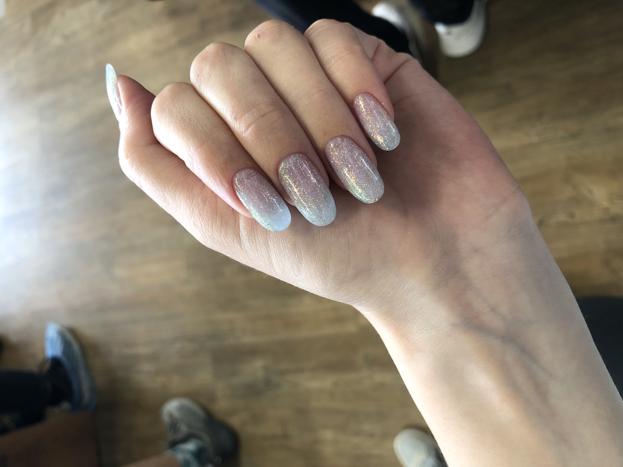 A person's left hand with five fingernails painted in a sparkly, ombre style, transitioning from a light blue or white at the tips to a shimmering silver or iridescent shade towards the base of the nail, is in focus. The nails are oval or almond-shaped and moderately long. The hand appears fair-skinned, with visible veins on the wrist and forearm. The background shows a blurred interior scene with people wearing shoes. The flooring appears to be made of wooden planks, positioned at an angle from upper left to lower right. There is no definitive evidence to suggest that the location of the image is Chaikovsky, Russia.